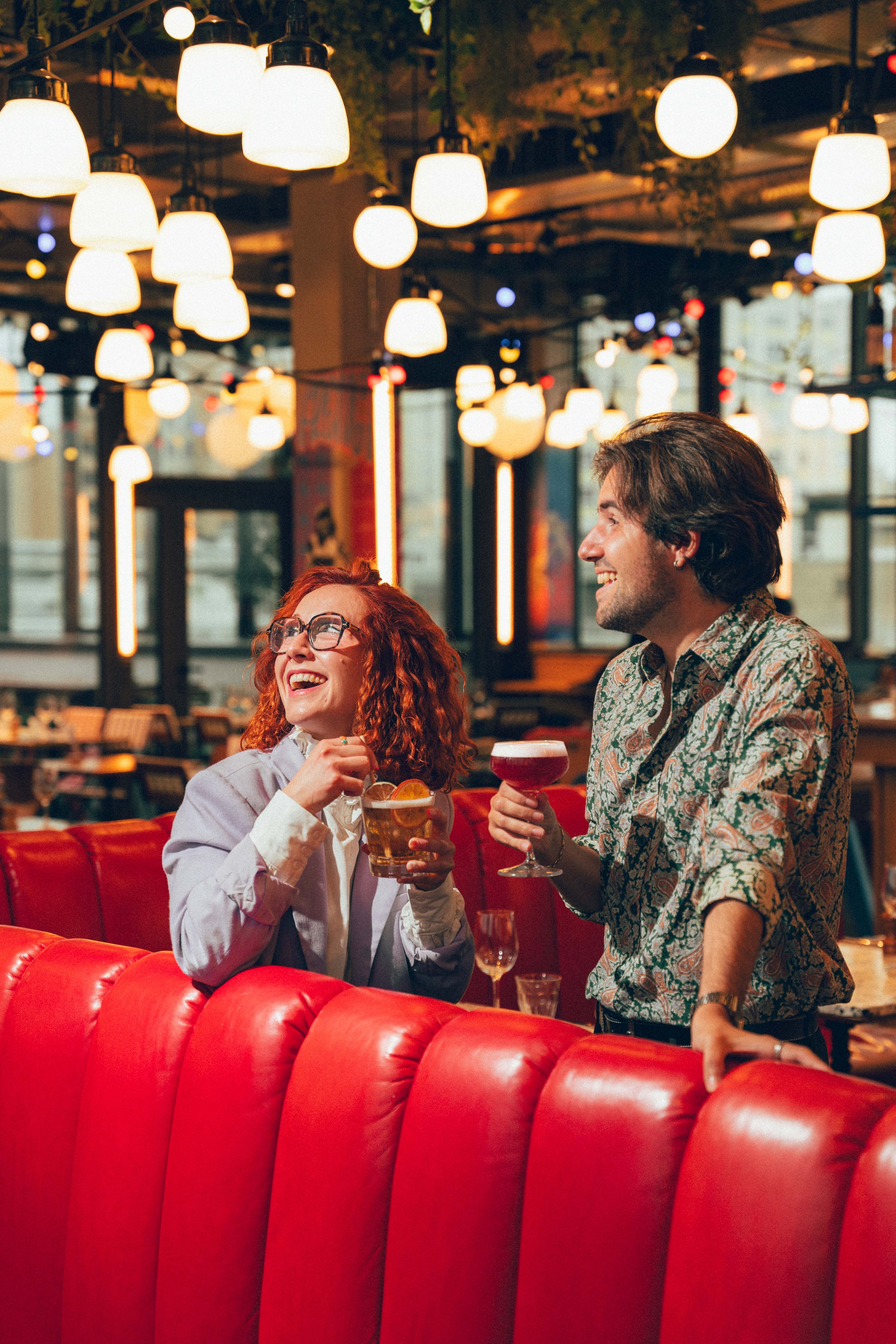 Deux personnes souriantes, une femme avec des cheveux roux et des lunettes, une homme avec des cheveux bruns longs, tenant chacune un verre, dans un restaurant ou bar avec un décor chaleureux et un éclairage d'ambiance