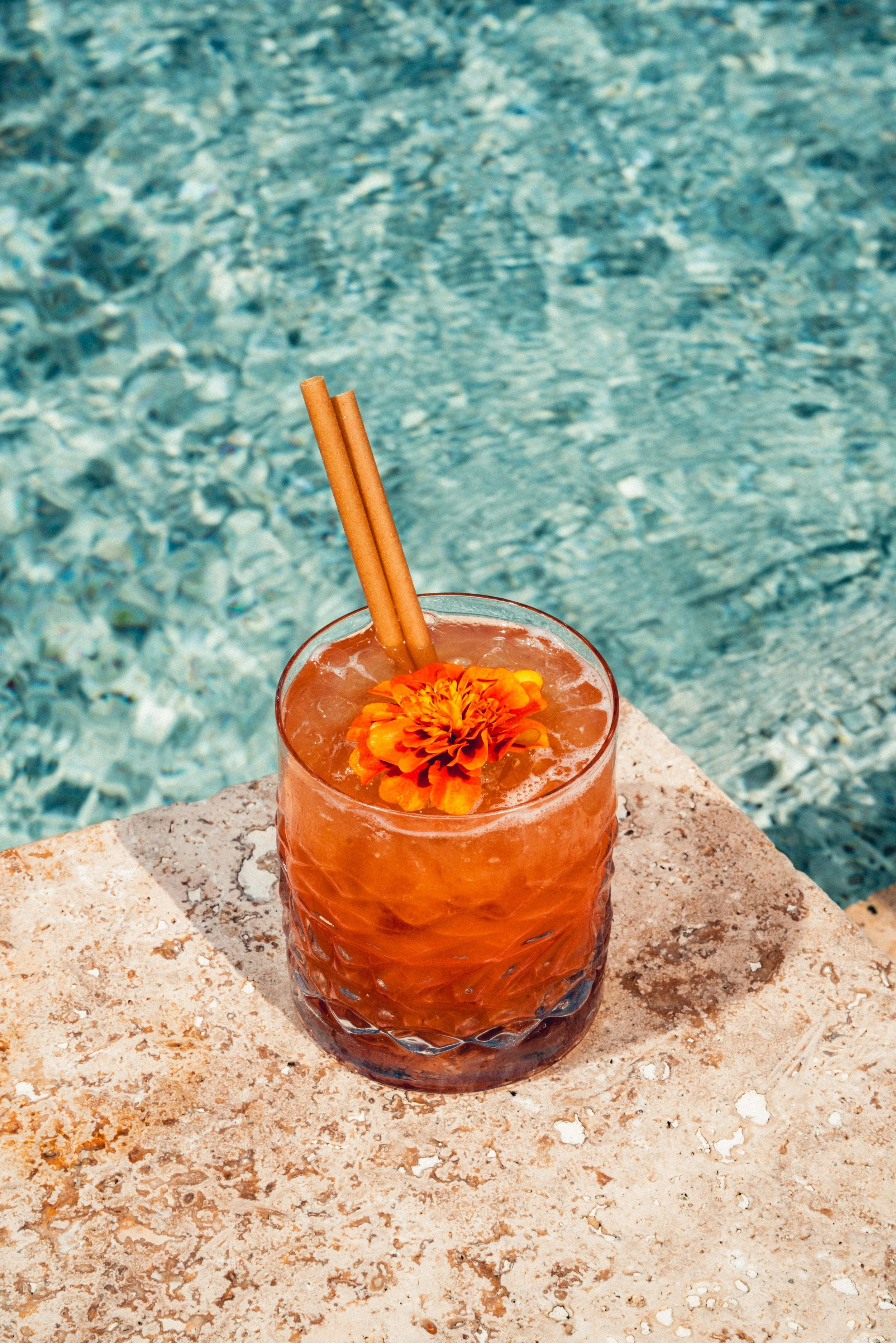 Cocktail orange dans un verre avec une fleur orange en décoration, deux bâtons de cannelle, sur une surface en pierre à côté d'une piscine d'eau turquoise.
