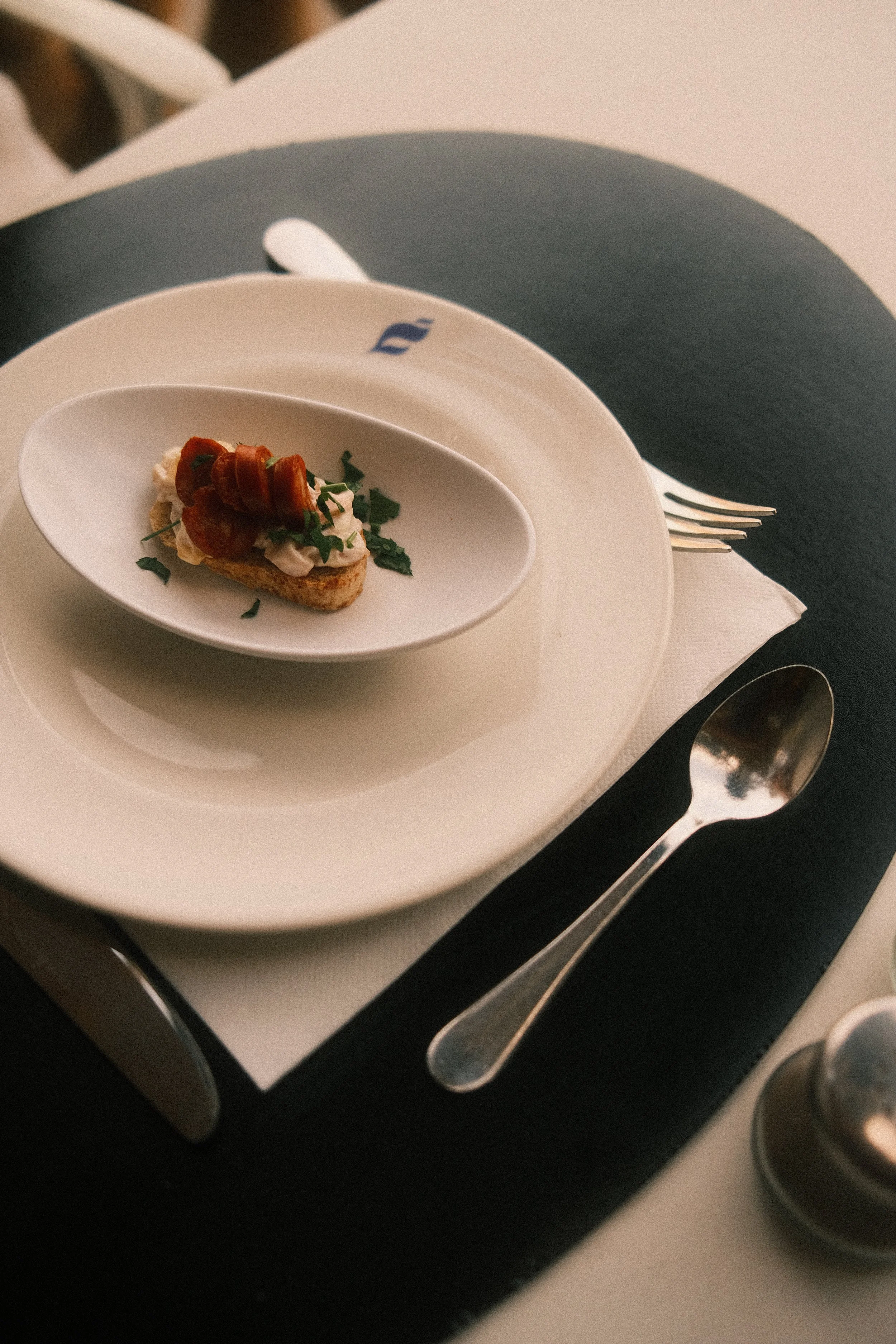 Une assiette de toast garnie de crème, de jambon, de feuilles de persil et de tranches de tomates cerises, sur une table avec une fourchette et une cuillère.
