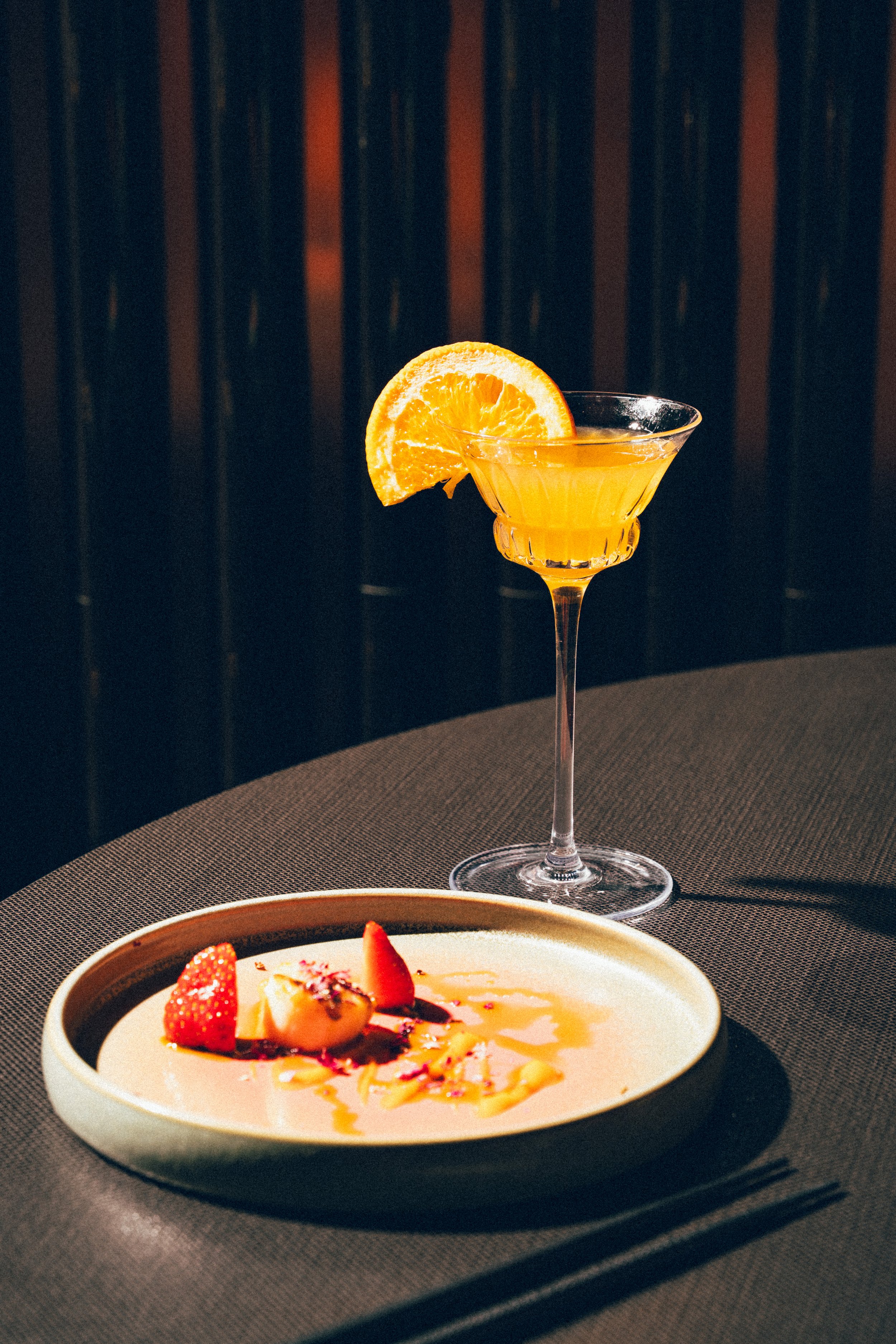 Un cocktail orange garni d'une tranche d'orange citron dans un verre à martini, et un plat de dessert avec des fraises et des fruits, sur une table sombre.