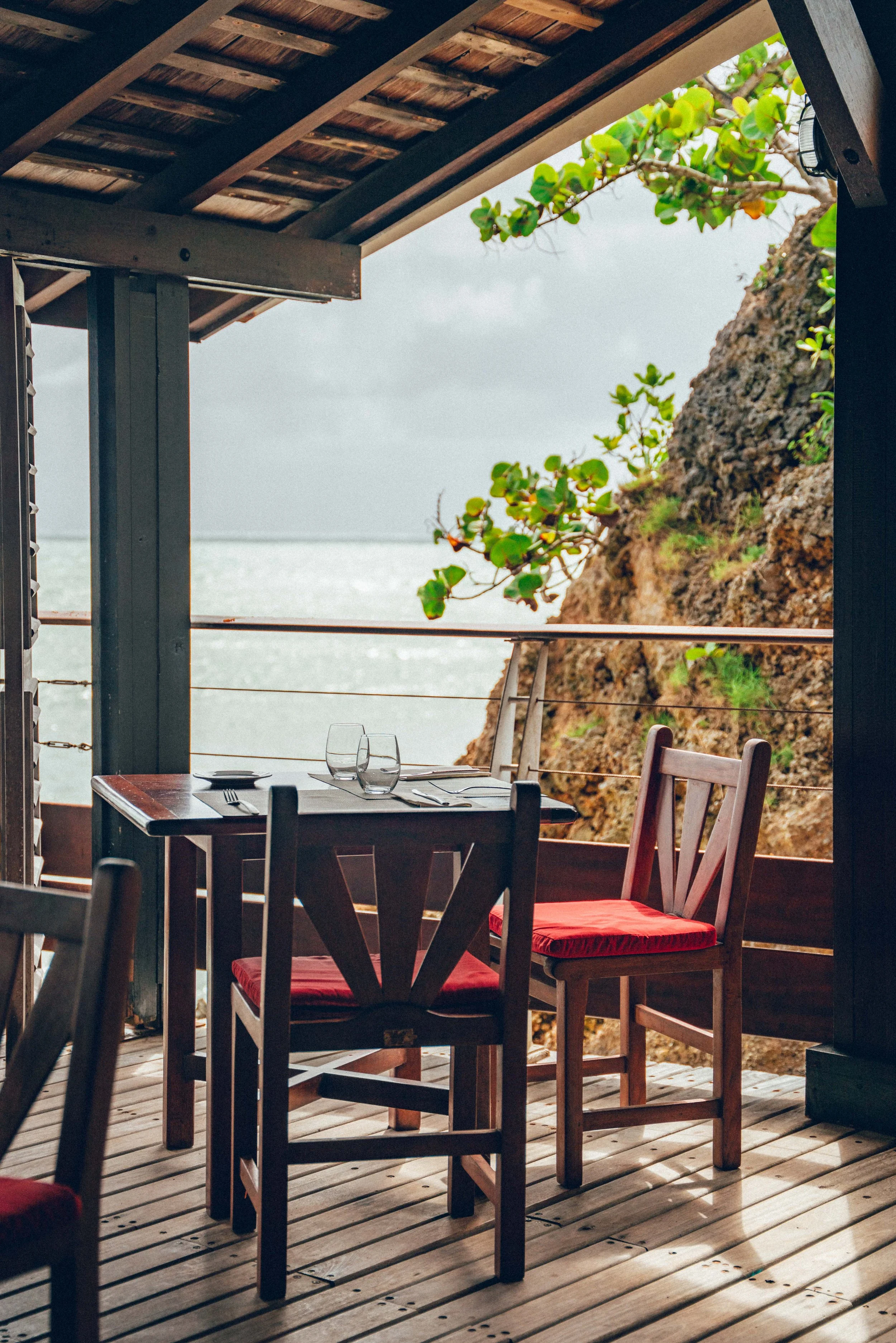 Table de restaurant avec deux verres, assiettes, couverts et chaises en bois avec coussins rouges, vue sur la mer et une falaise rocheuse dans un espace en plein air sous un toit en bois.