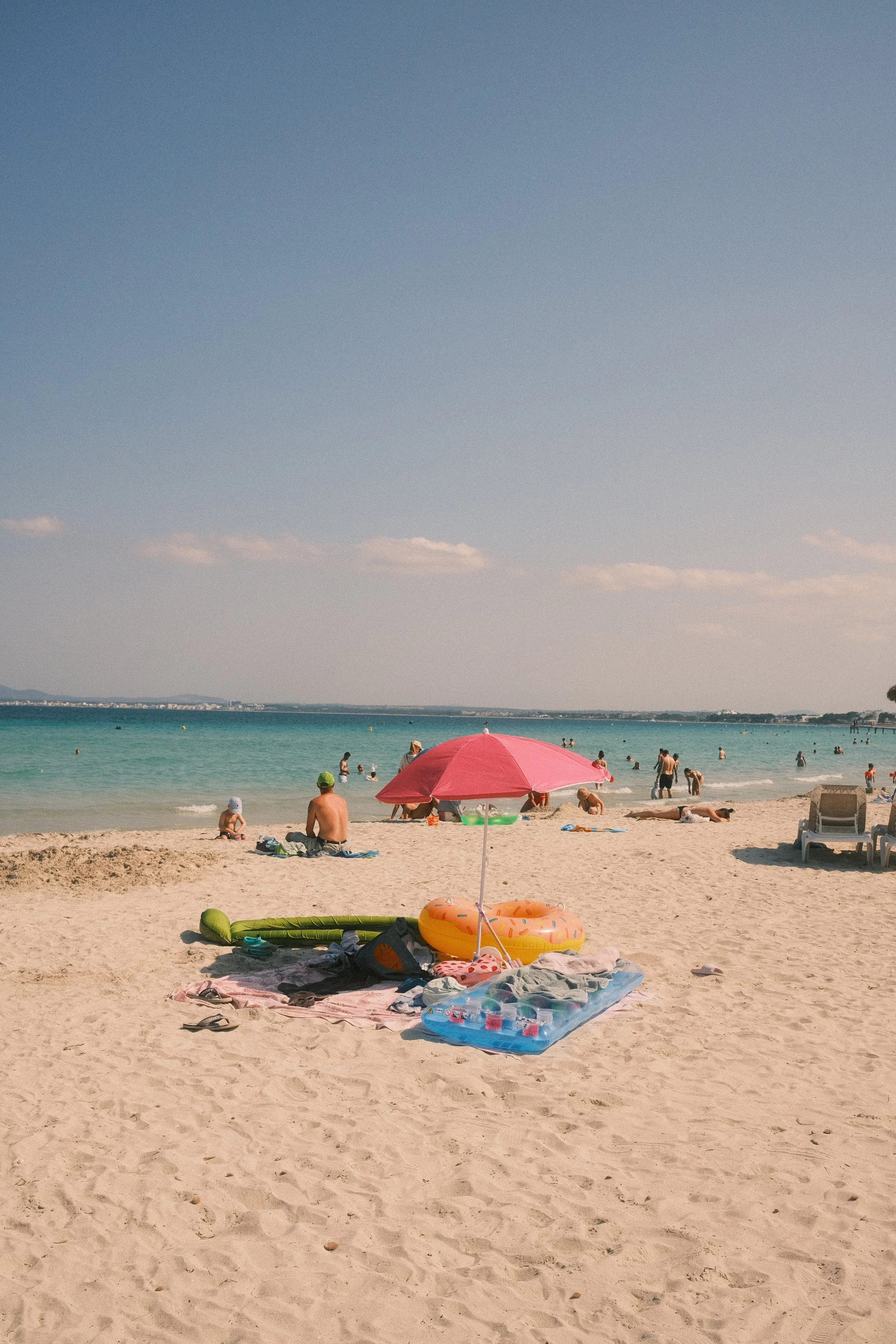 Plage ensoleillée avec des personnes nageant dans la mer, un parasol rose, des tapis et jouets sur le sable, un ciel clair avec quelques nuages, et une vue lointaine sur la côte et la ville.