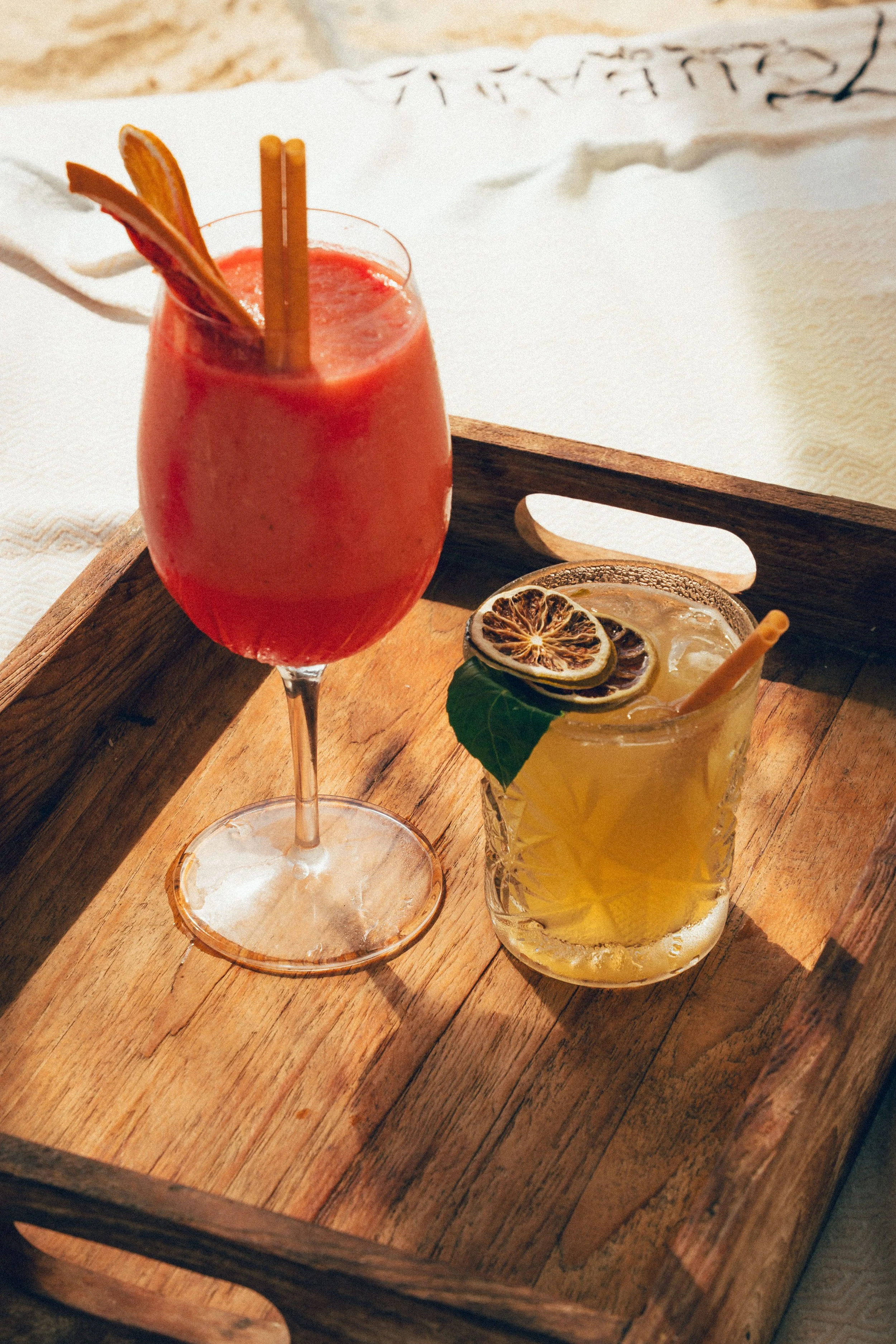 Deux cocktails colorés, un rouge dans un verre à vin et un jaune dans un verre en mason jar, garnis de tranches de citron ou d'orange et de feuilles de menthe. Servis sur une table en bois avec un fond clair.
