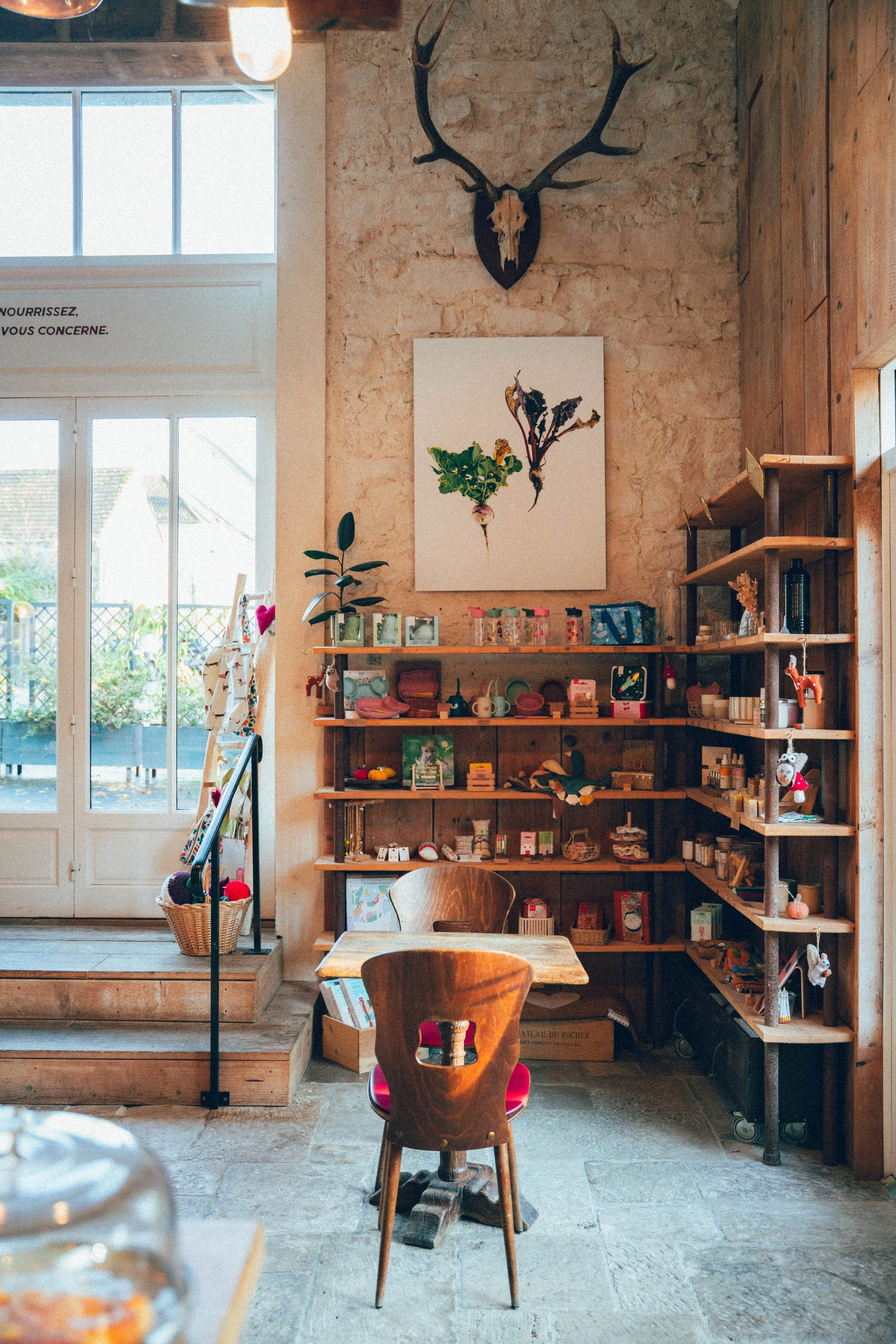 Intérieur d'une boutique ou d'un café avec un mur en briques, une étagère en bois remplie d'objets, une table en bois avec une chaise en bois, un mur en pierre, et une décoration de tête de cerf avec des cornes suspendue au mur.