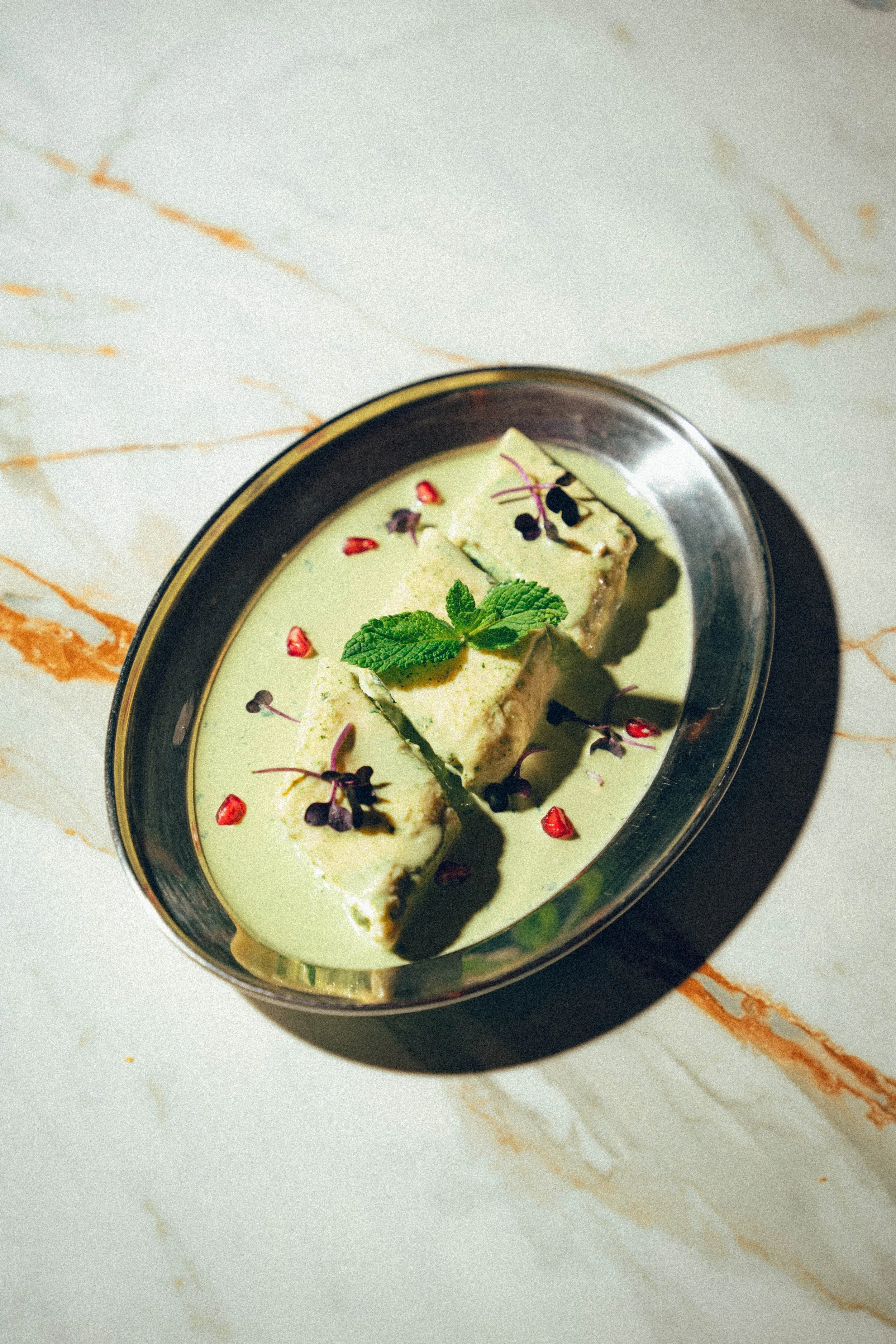 Un plat de dessert avec une mousse verte, décoré de feuilles de menthe, des grains de grenade, des micro-pousses, dans un bol en céramique