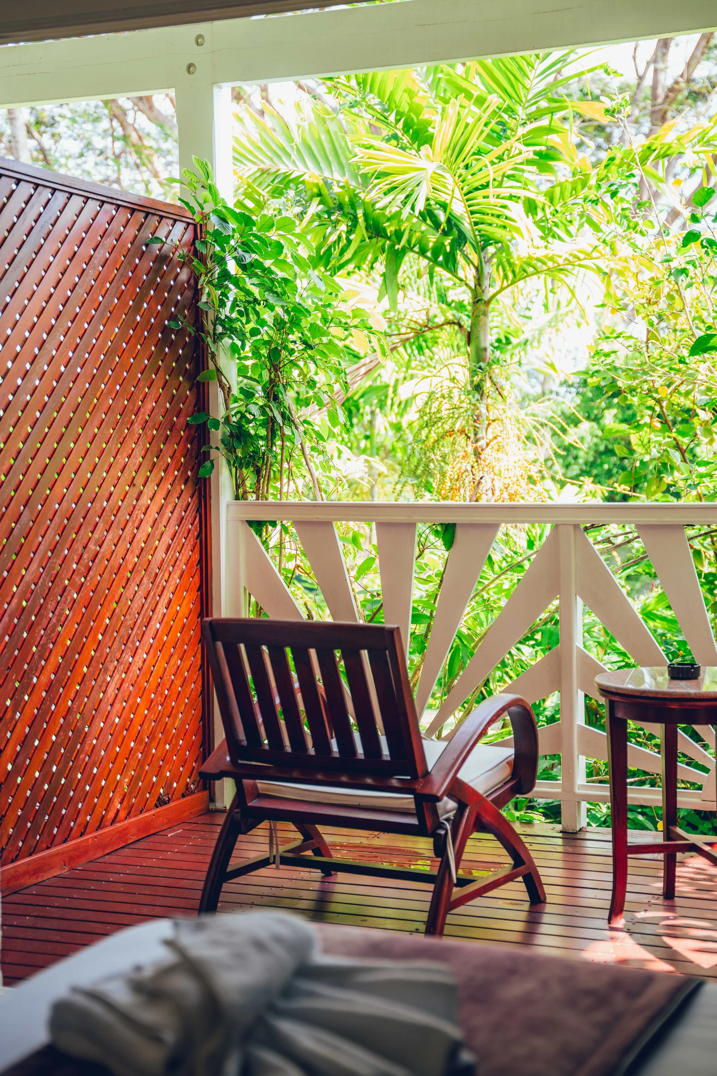 Photographie hôtelière d'une chambre élégante dans un hôtel haut de gamme. Balcon avec chaises en bois, vue sur un jardin avec des grandes plantes vertes et des arbres en arrière-plan, lumière du soleil.