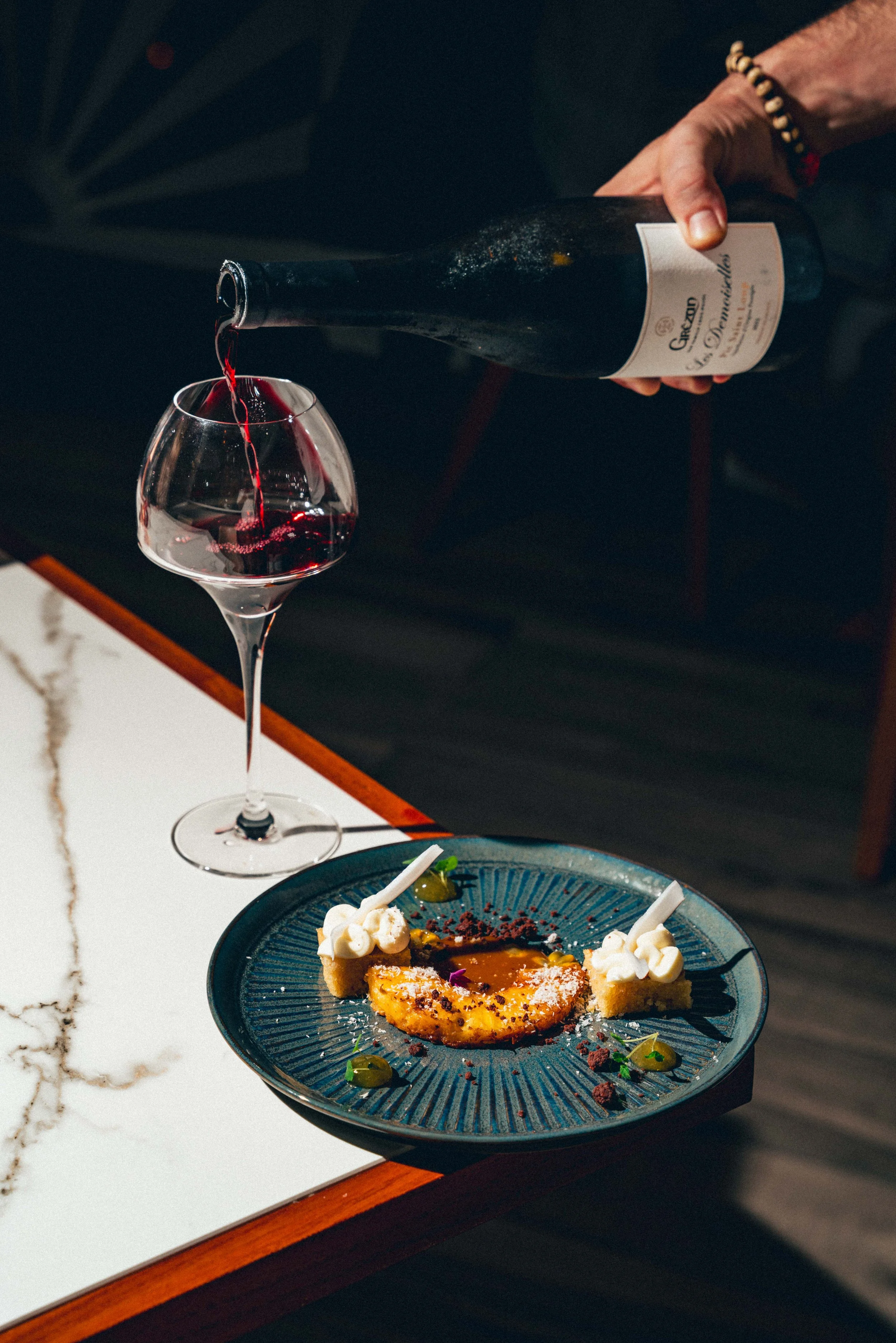 Photographie culinaire d'une personne versant du vin rouge dans un verre, à côté d'une assiette avec un dessert décoré, sur une table en marbre avec une surface en bois visible.