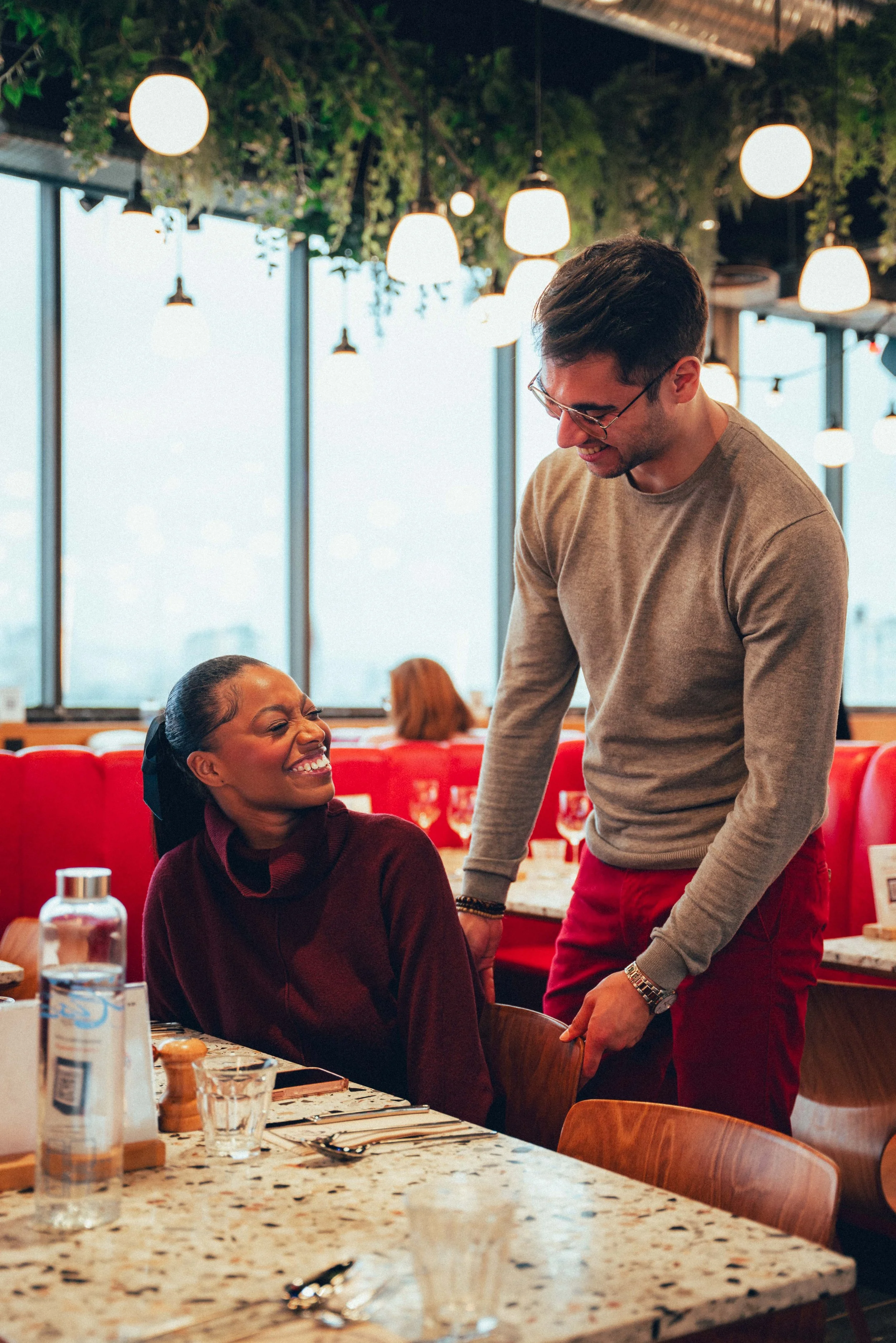 Un homme souriant parle avec une femme assise à une table dans un restaurant, ambiance chaleureuse avec éclairage suspendu et décoration végétale.