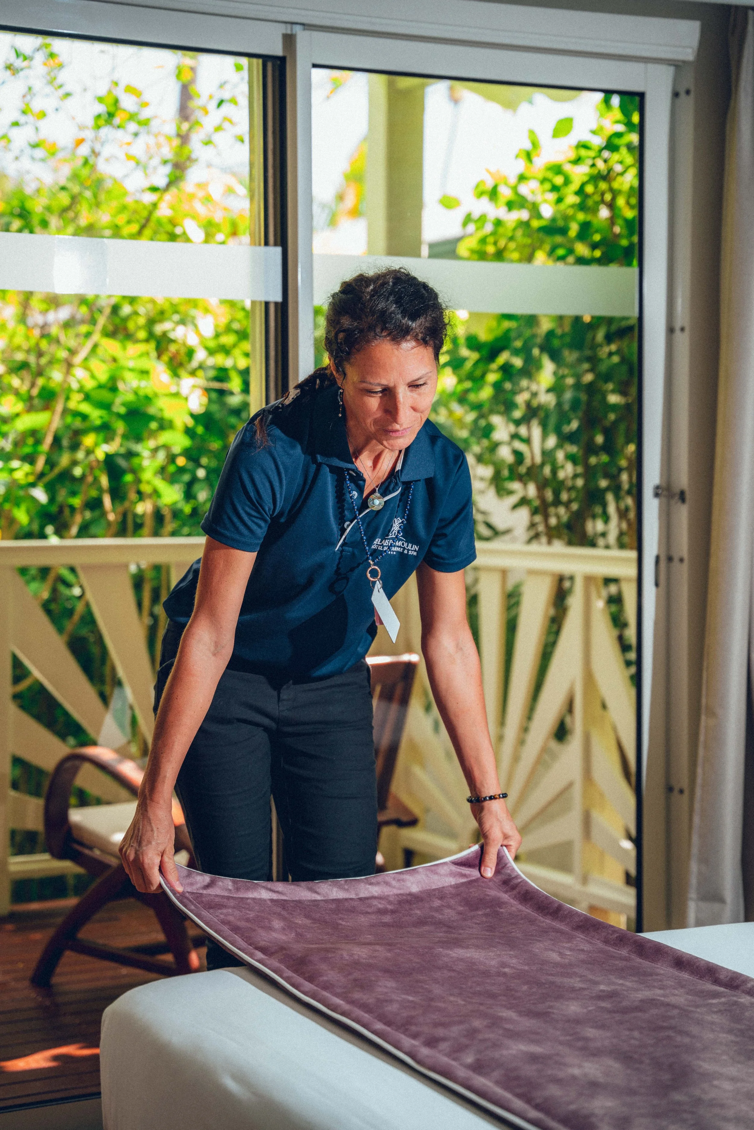Une femme âgée plie une nappe en tissu violet à l'intérieur d'une pièce lumineuse avec vue sur un jardin vert par la fenêtre.