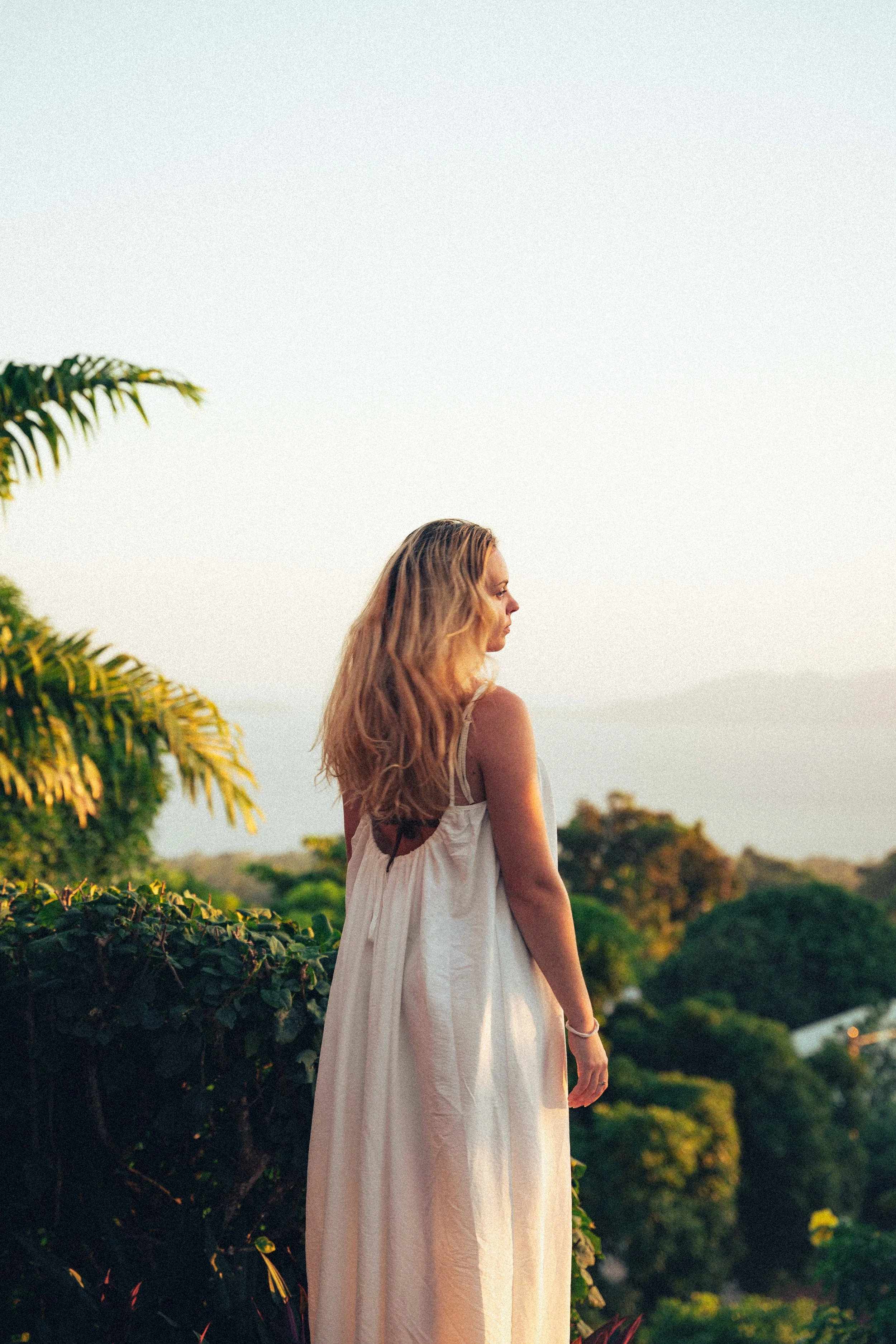 Femme aux cheveux longs et bouclés portant une robe blanche, debout dans un jardin avec des arbres et un ciel clair.