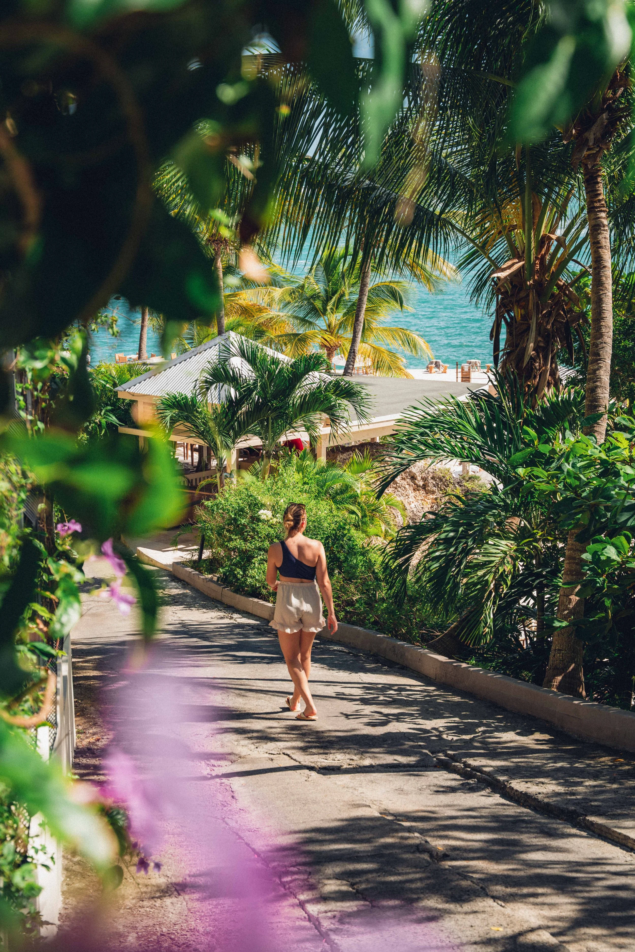 Une femme marche sur un chemin entouré de végétation tropicale, avec vue sur la mer en arrière-plan.