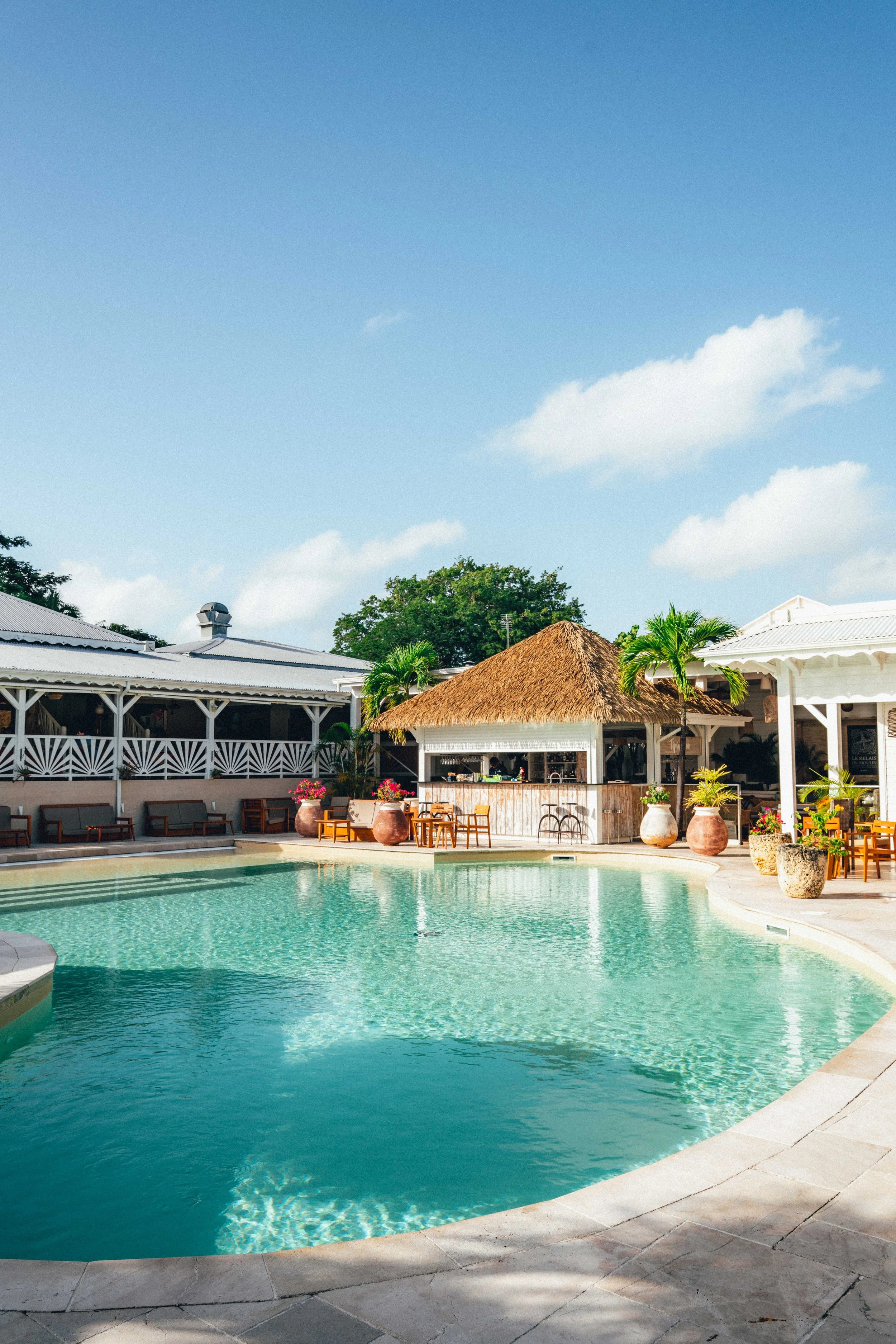 Photographie hôtelière d'une piscine extérieure avec eau turquoise, entourée de chaises longues et de grands pots avec des plantes, bâtiments en style côtier avec toits en chaume et en métal, arbres et ciel bleu avec quelques nuages