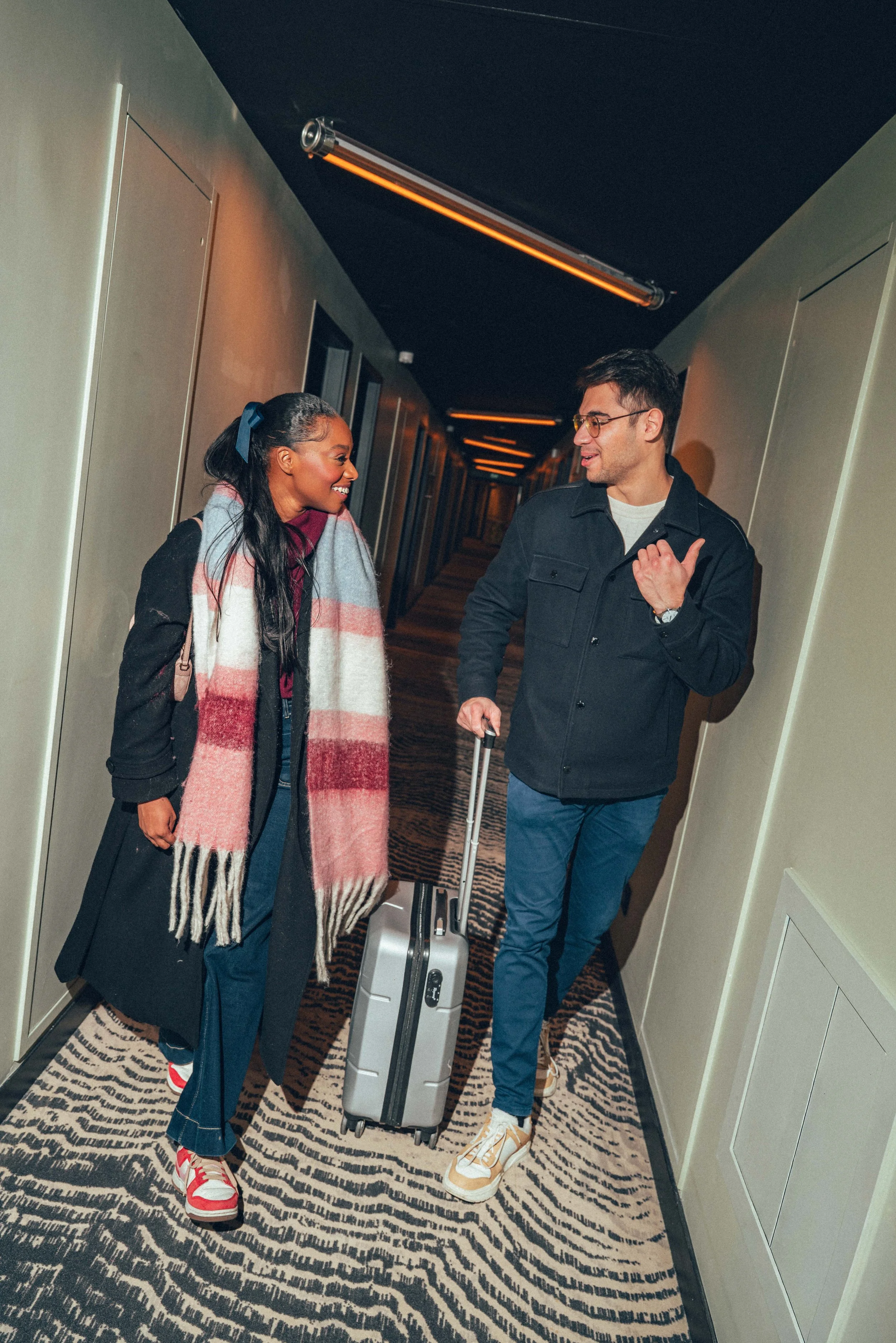Deux personnes discutent dans un couloir d'hôtel, l'une avec une valise à roulettes. Ils semblent être titulaires, souriants et engagés dans une conversation amicale.