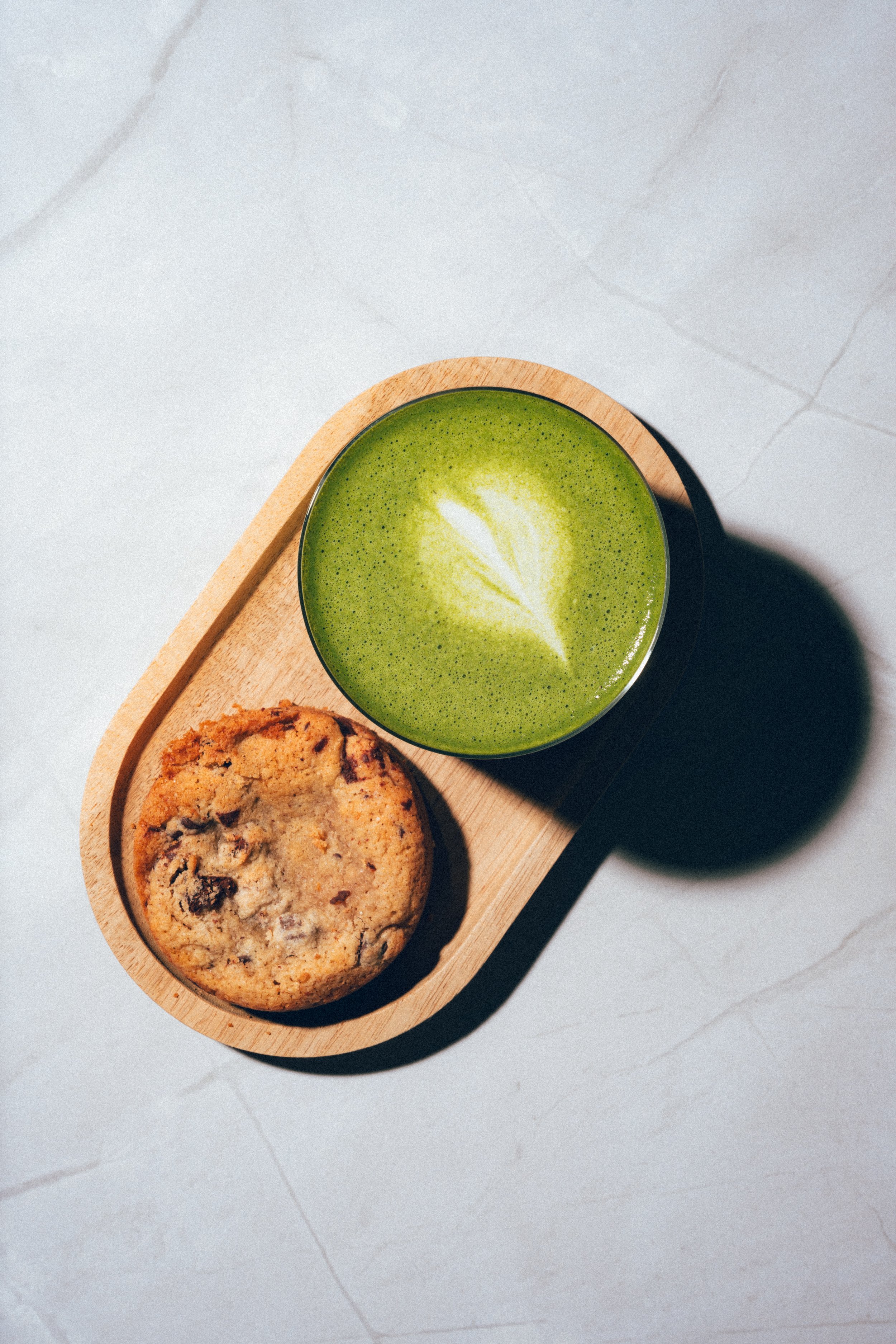 Un plateau en bois contenant un biscuit aux pépites de chocolat et une boisson verte avec un motif de feuille dessiné dedans, posé sur une surface en marbre clair.