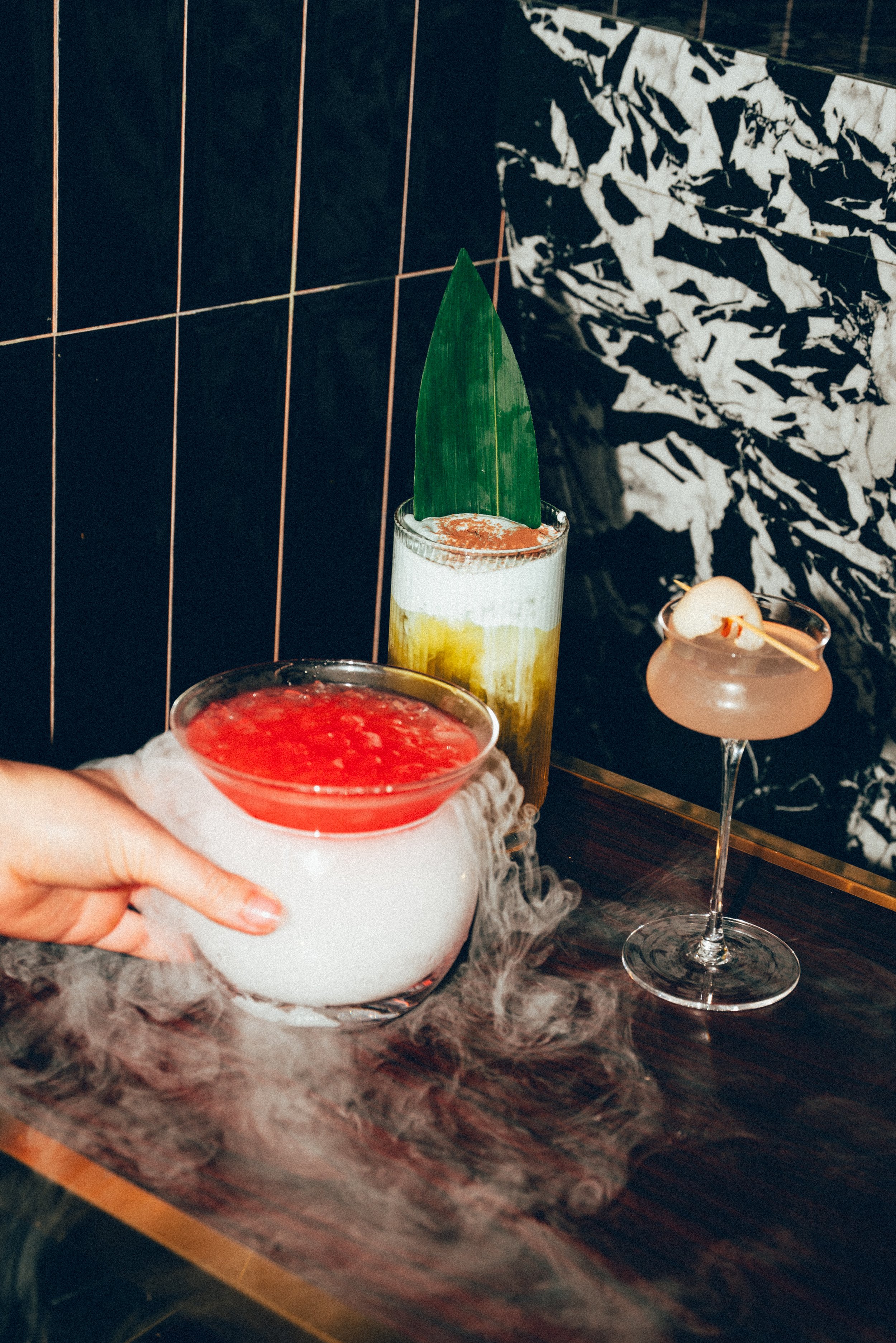 Plusieurs cocktails colorés sur un bar en marbre, dont un avec une feuille de pandan, un autre avec une cerise, et un verre fumant avec une boisson rose, dans un décor bar élégant.