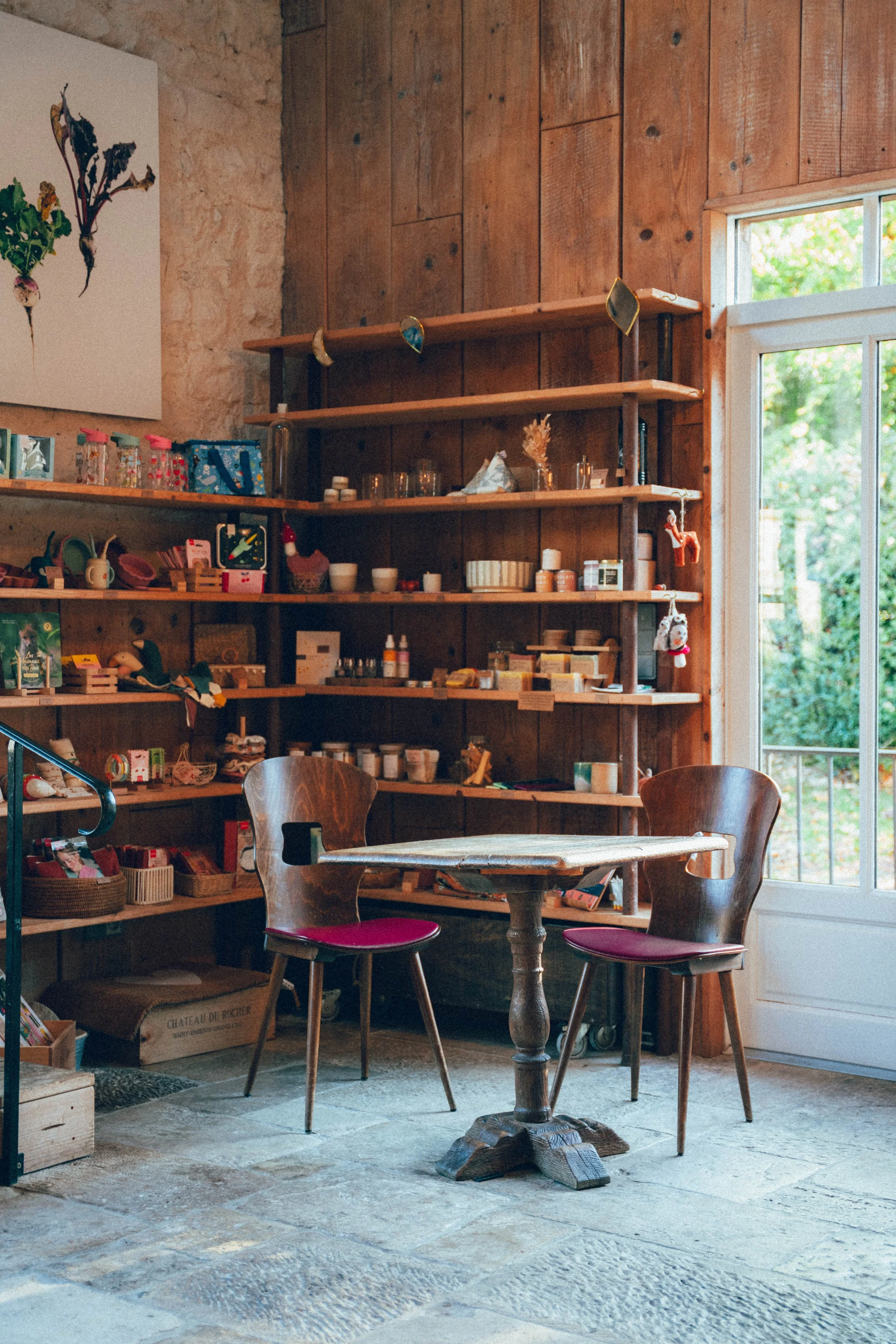 Une salle avec des étagères en bois remplies d'objets divers, un mur en pierre et en bois, une grande fenêtre laissant entrer la lumière naturelle, et une table ronde avec deux chaises en bois avec des coussins rouges.
