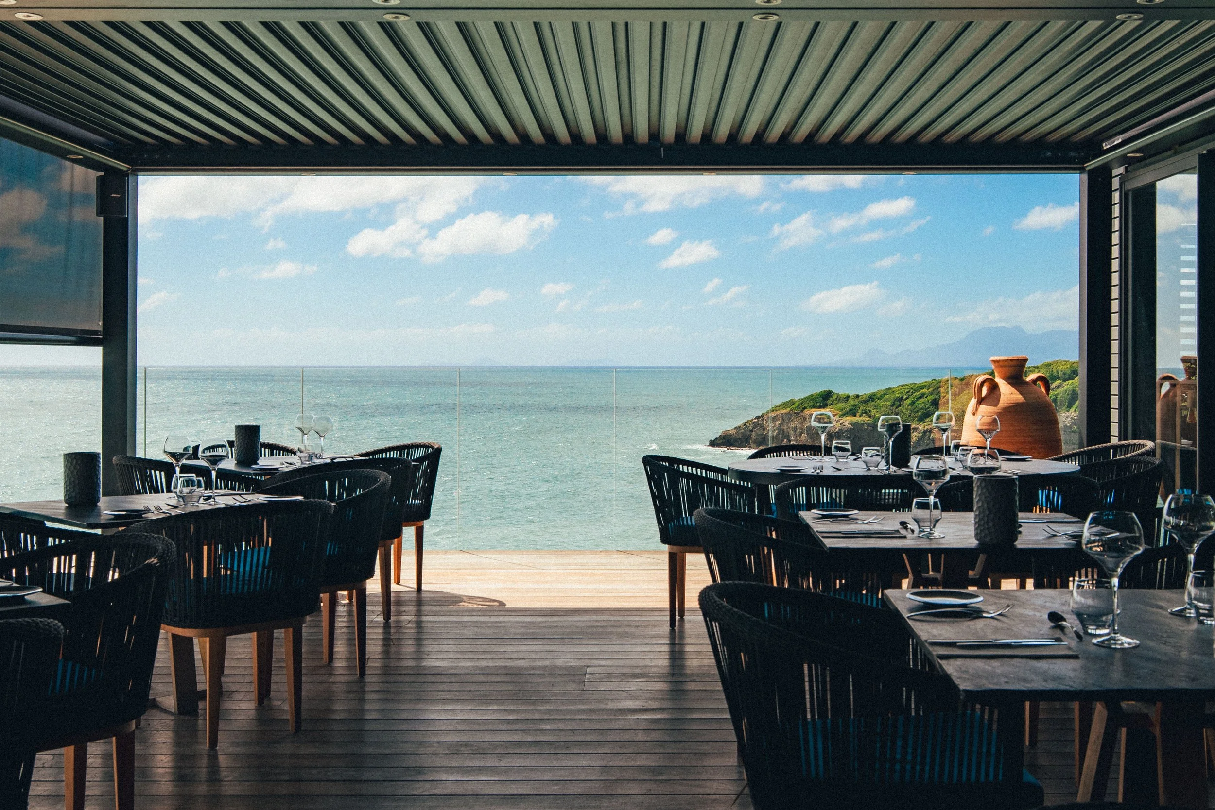 Salle à manger avec vue sur la mer, tables prêtes pour le service, décor moderne, vue panoramique sur un ciel en partie nuageux et des collines au loin.