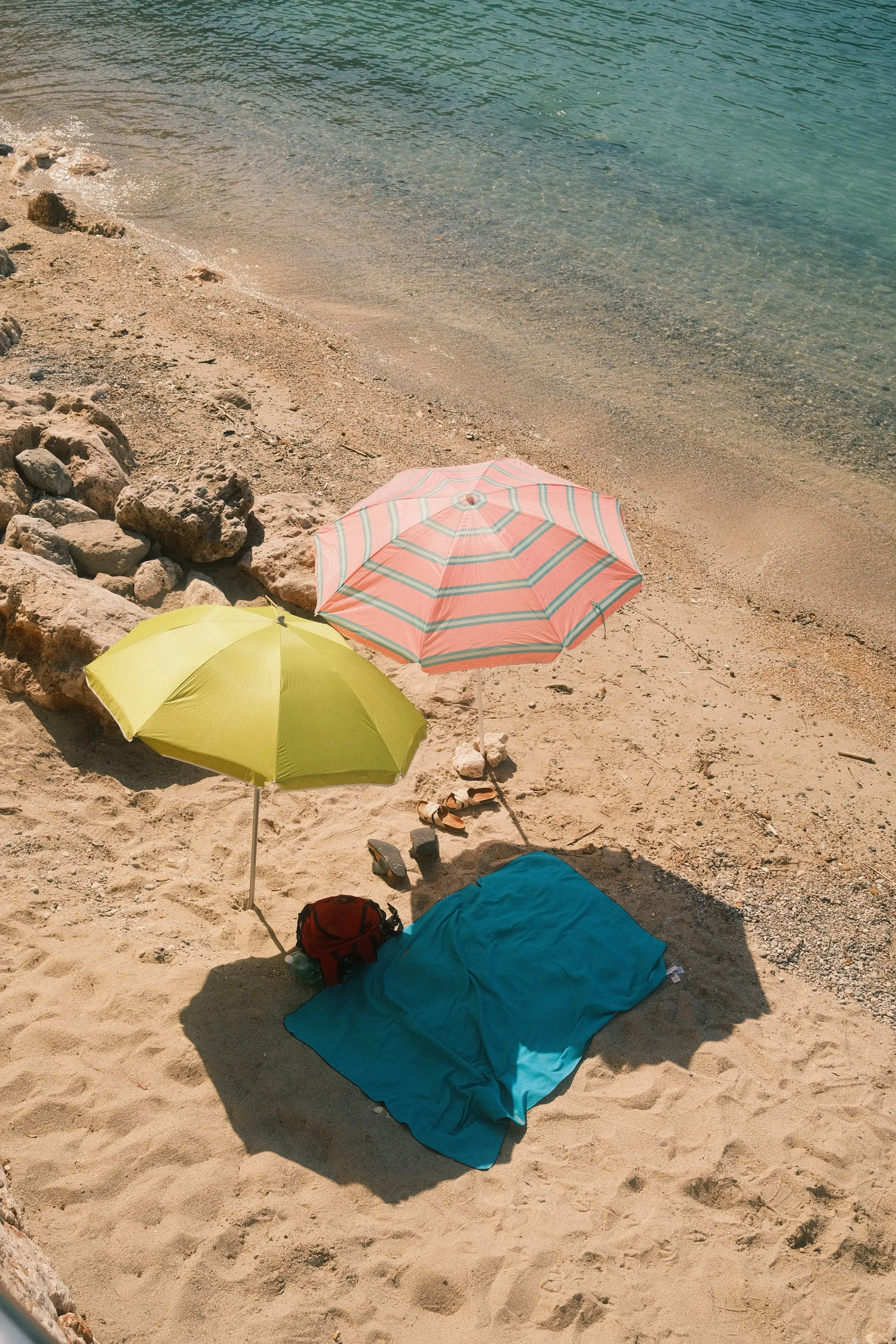 Plage avec sable, rochers, parasols, serviette bleue, sacs à dos, chaussures, et proches de l'eau turquoise.