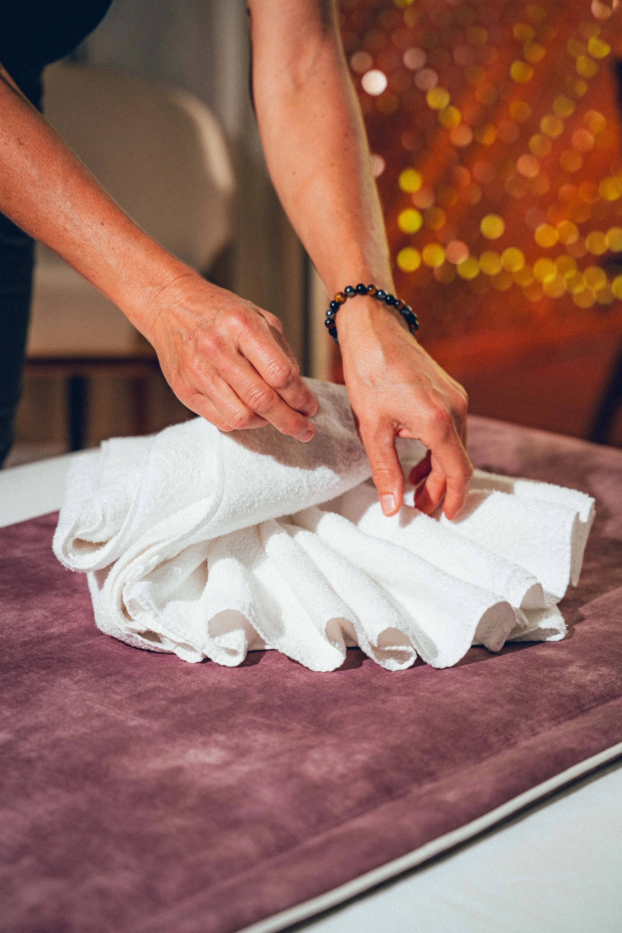Une personne plie une serviette blanche sur une table recouverte d'un tapis violet. La personne porte un bracelet à perles noires et une robe noire. En arrière-plan, il y a des lumières floues de couleur jaune et rouge.