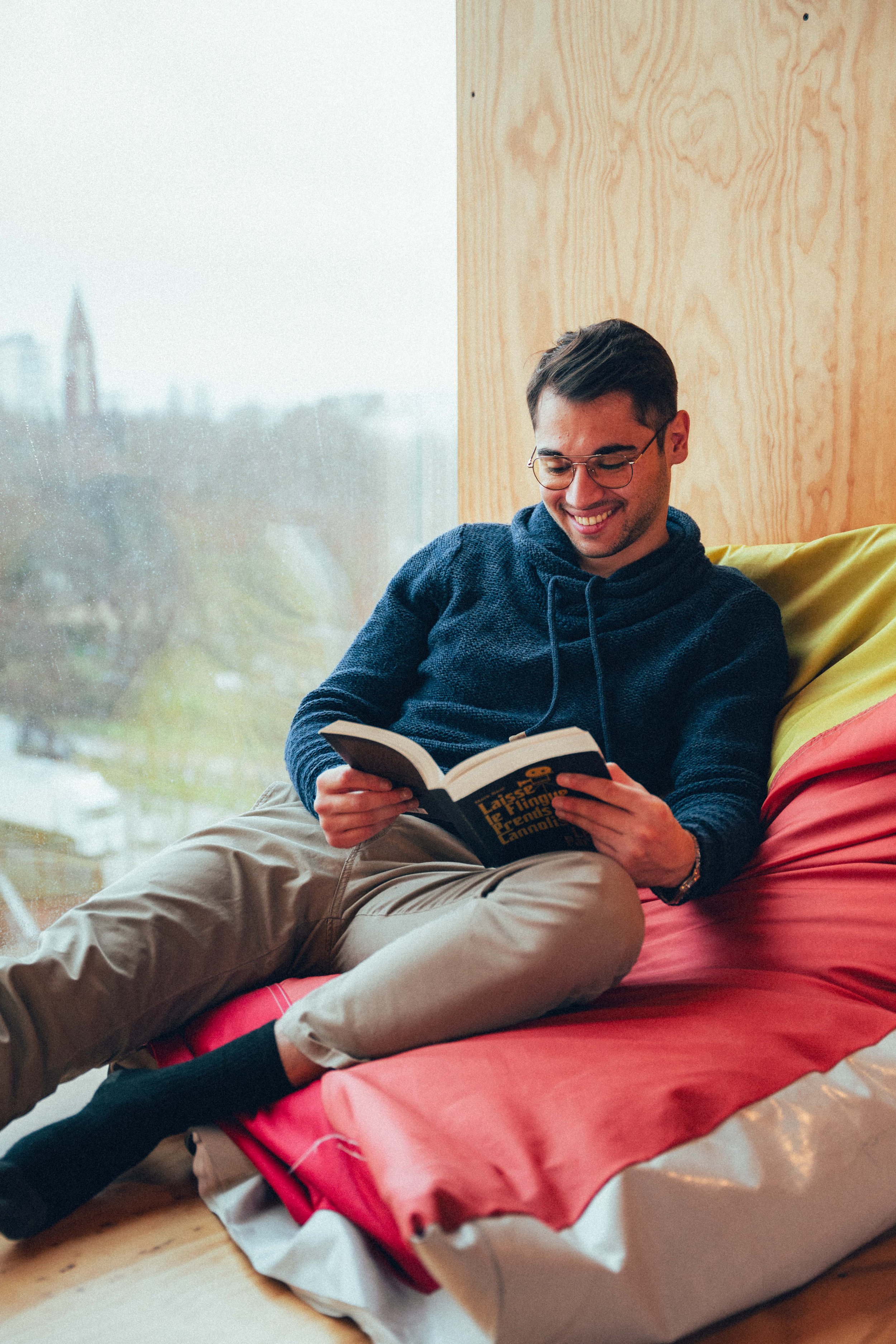 Jeune homme souriant, portant lunettes, assis sur un matelas rose, lisant un livre dans une pièce avec une fenêtreLarge vue de l'intérieur d'une pièce lumineuse avec un homme qui lit, assis sur un tapis avec un mur en bois derrière lui et une grande 