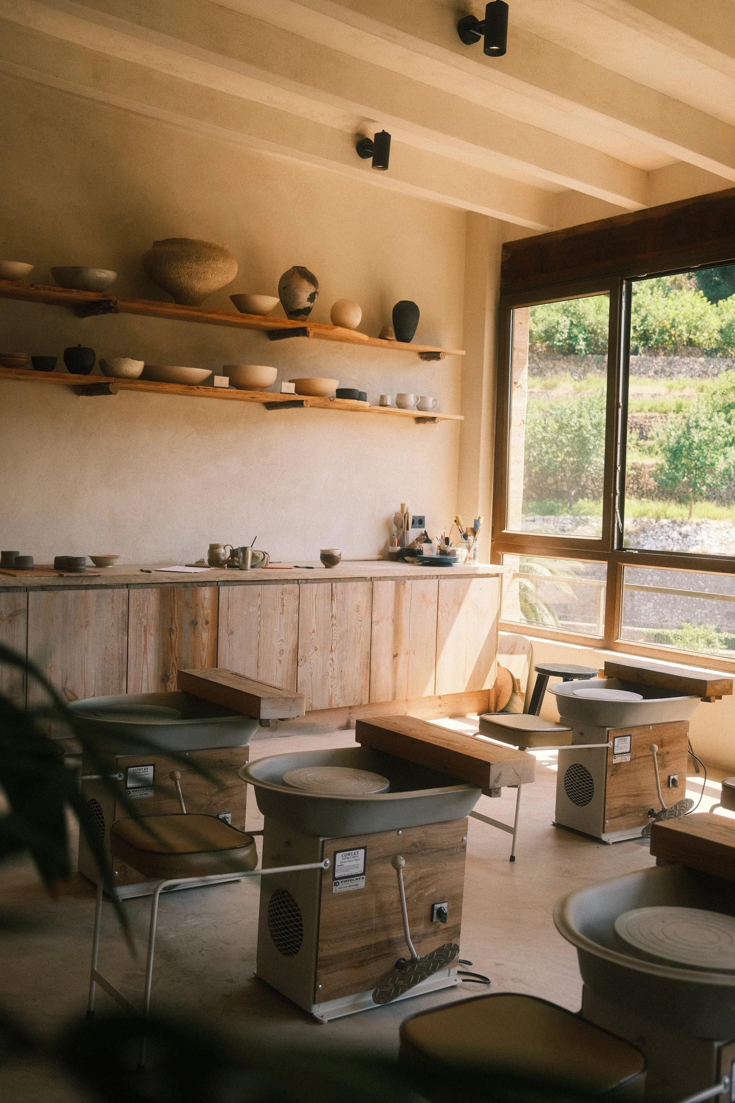 Une salle d'atelier de poterie avec des tours de poterie en bois, des céramiques en cours de fabrication et une grande fenêtre laissant entrer la lumière naturelle sur un paysage verdoyant dehors.