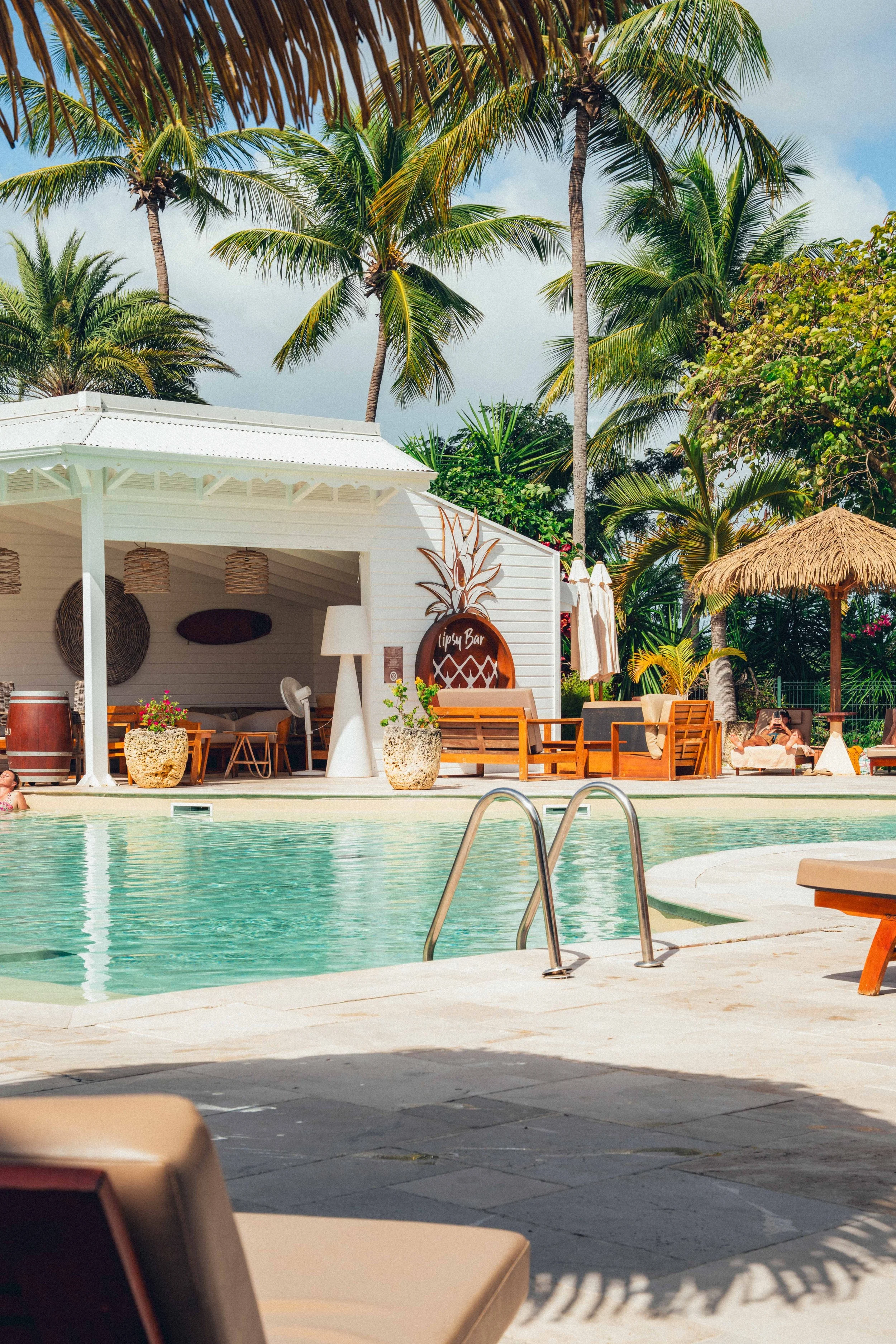 Photographie hôtelière d'une piscine avec des chaises longues en première plan, un bar de style tropical avec palmiers, hamacs et parasols en arrière-plan, dans un cadre tropical avec des palmiers et un ciel partiellement nuageux.