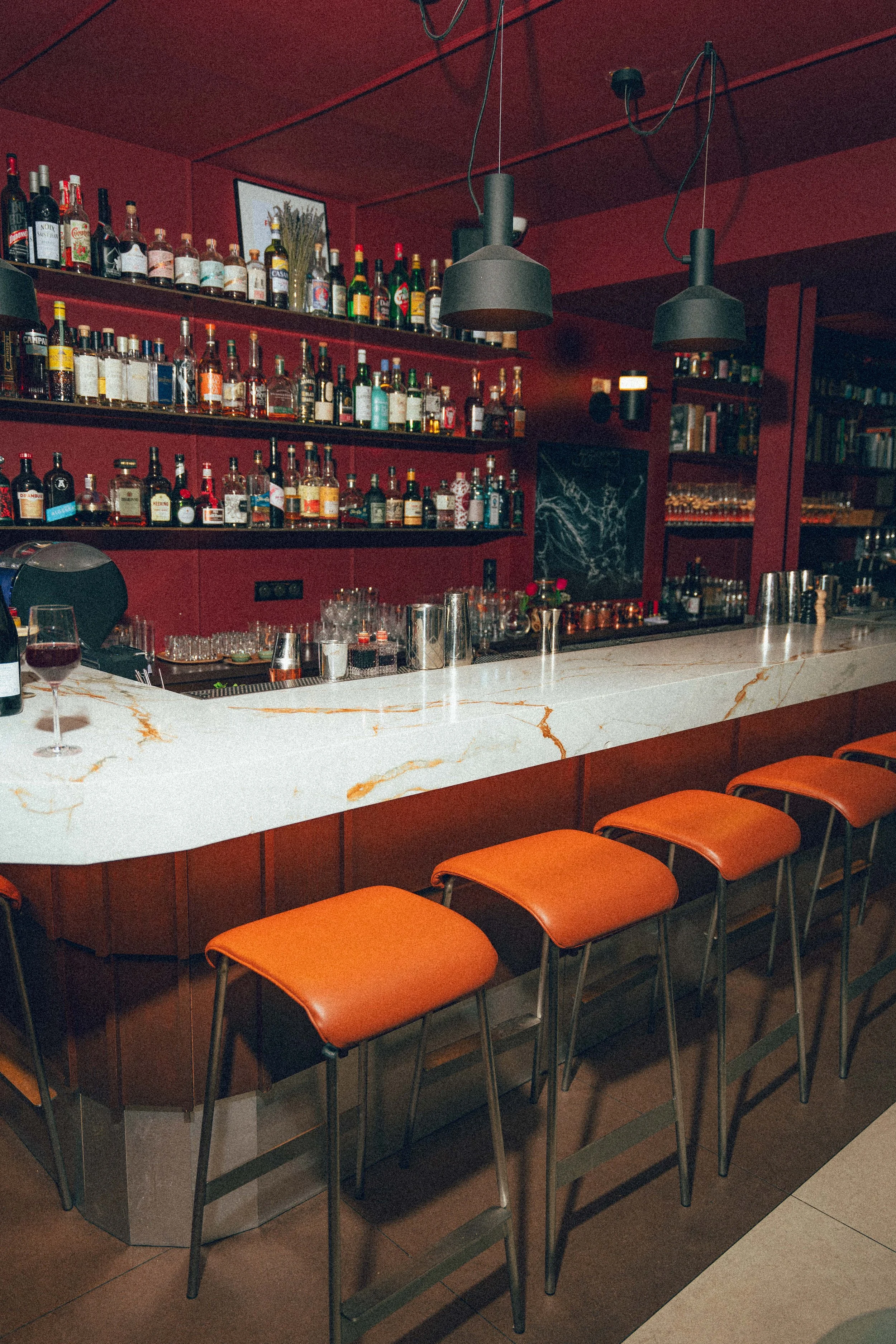 Bar avec étagères pleines de bouteilles d'alcool, comptoir en marbre blanc, chaises orange en face du comptoir, lampes suspendues noires, bibliothèque en arrière-plan.