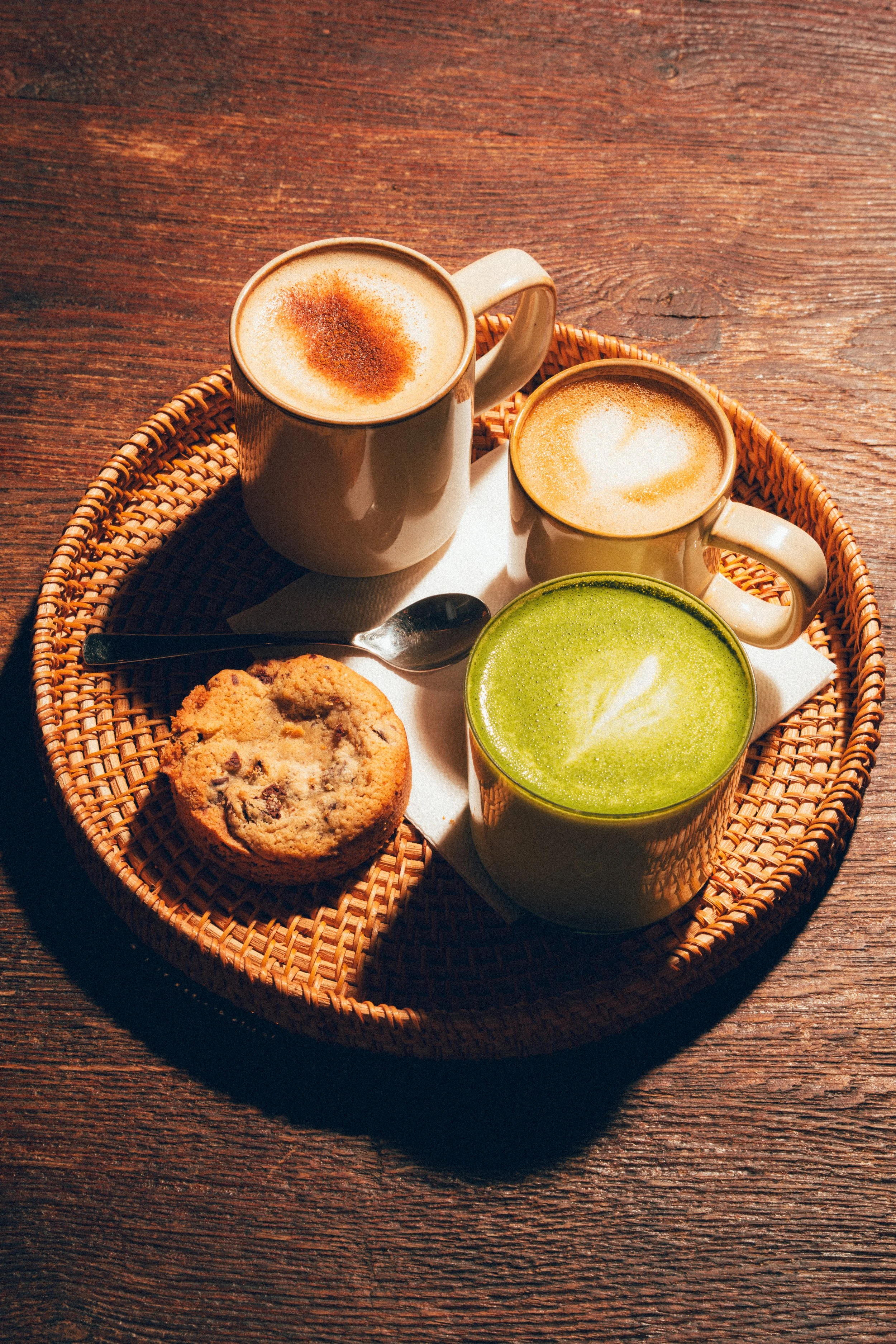 Trois tasses de café avec cappuccino, un cookie aux pépites de chocolat, et un smoothie vert dans un panier en rotin sur une table en bois.