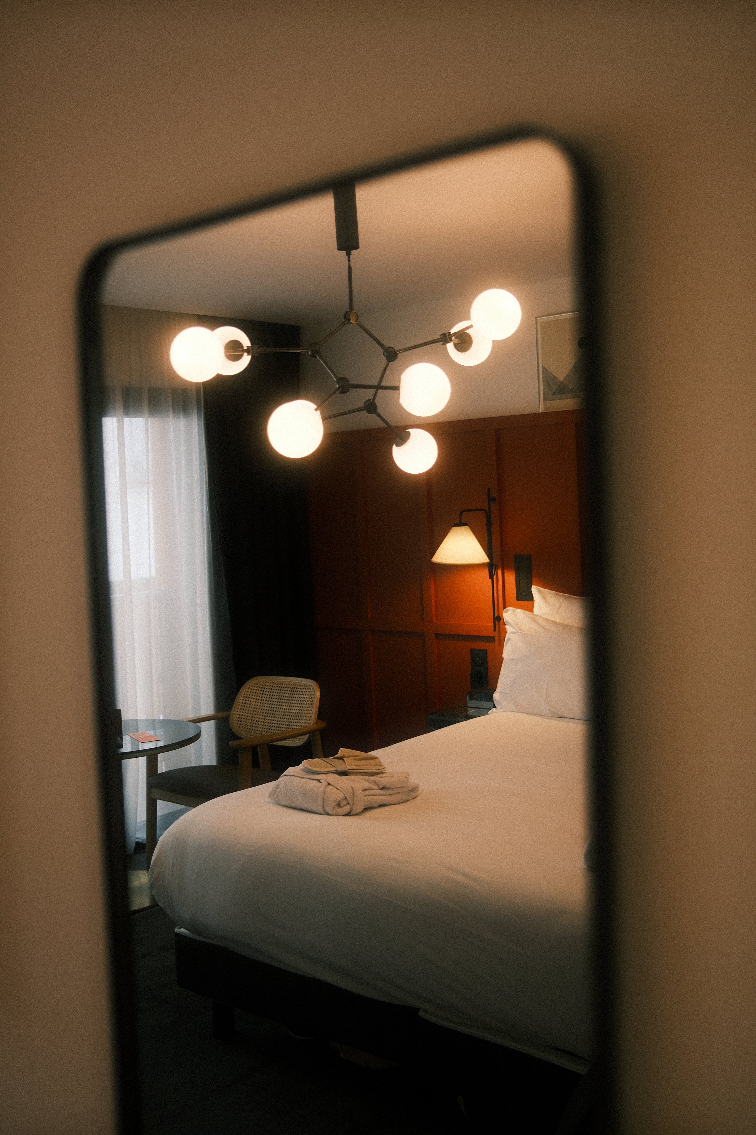 Reflet d'une chambre d'hôtel à travers un miroir, avec un lit fait, une lampe de chevet, un plafond moderne à plusieurs lumières, et un coin salon avec une table et quelques chaises.