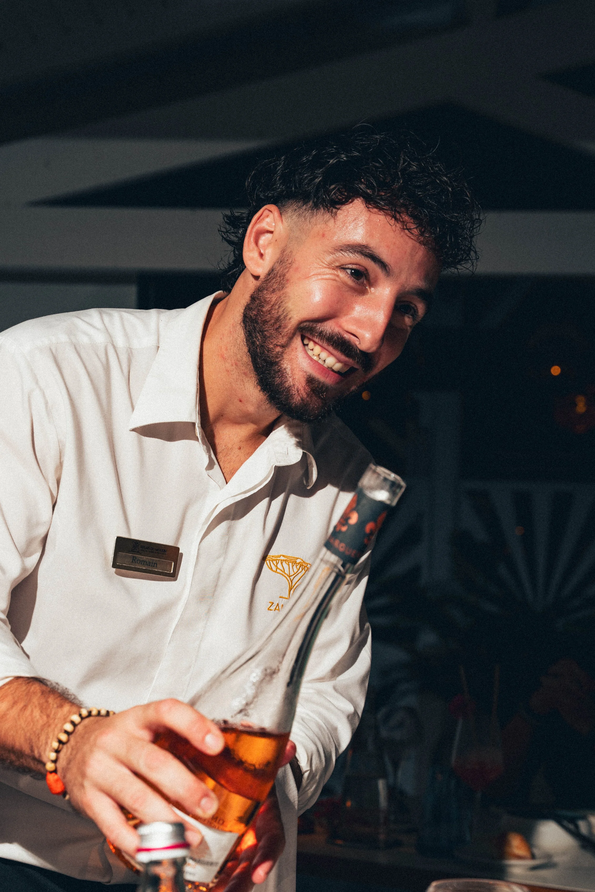 Un homme souriant portant une chemise blanche avec un badge "Romain" et une broche en forme de parasol, tenant une bière, dans un environnement intérieur.