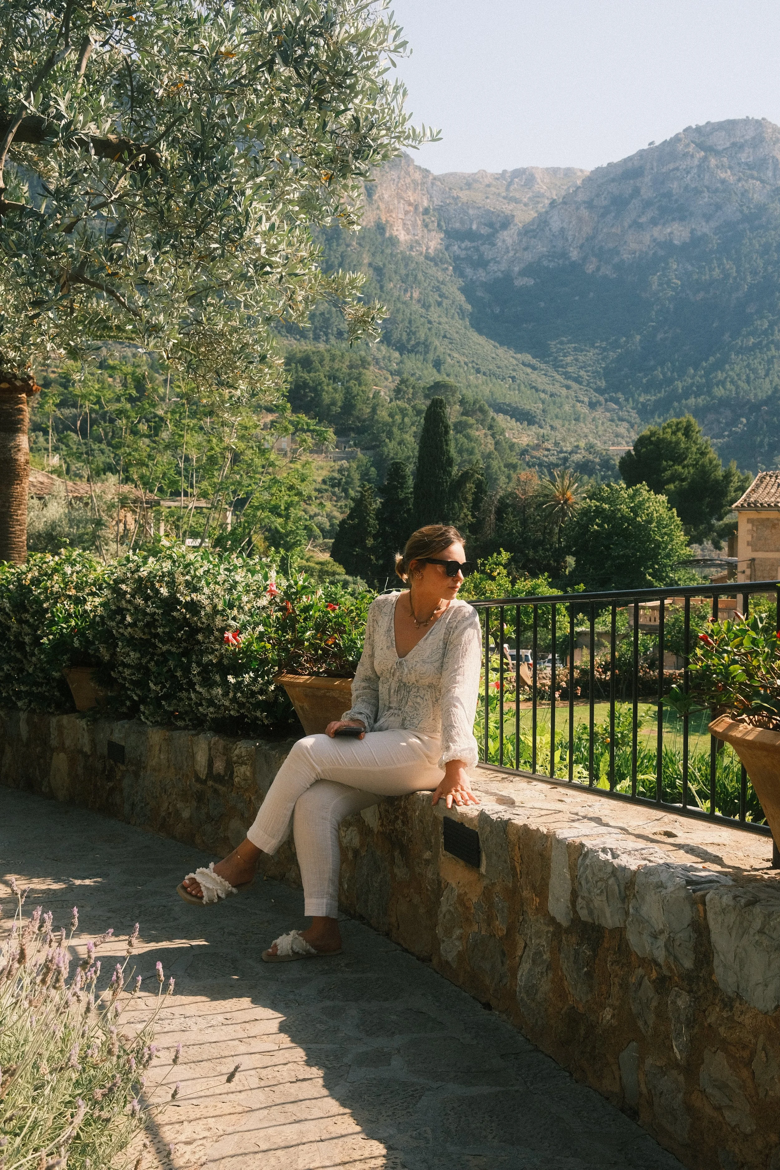 Une femme assise sur un muret en pierre dans un jardin, avec des montagnes en arrière-plan, portant des vêtements légers, lunettes de soleil, et des sandales, entourée de végétation verdoyante et de fleurs.