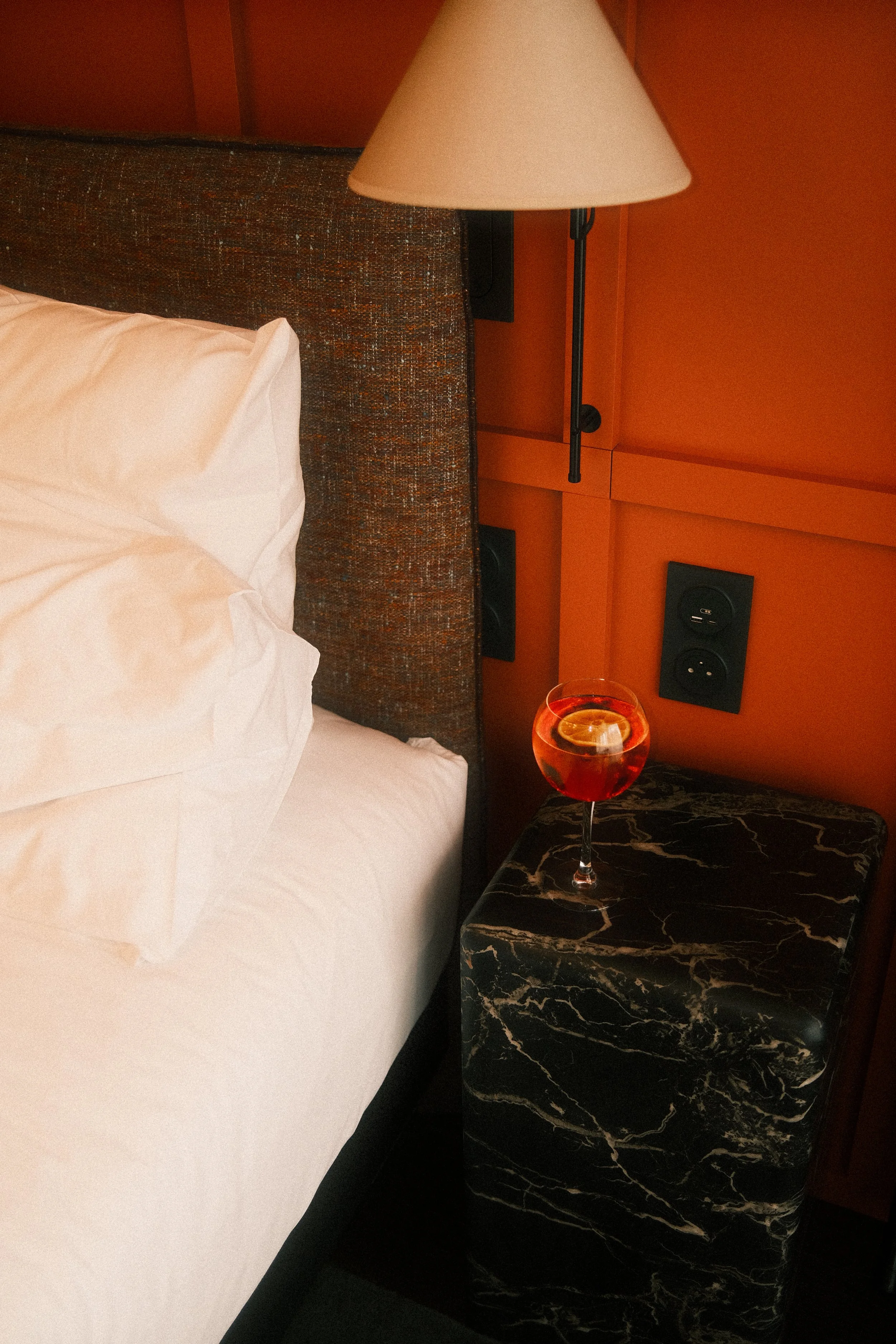 Une tête de lit rembourrée avec un tissu à motifs, une table de nuit en marbres noirs avec des veines dorées, une lampe de chevet, un verre de cocktail avec tranche de citron, et des prises électriques sur le mur orange.