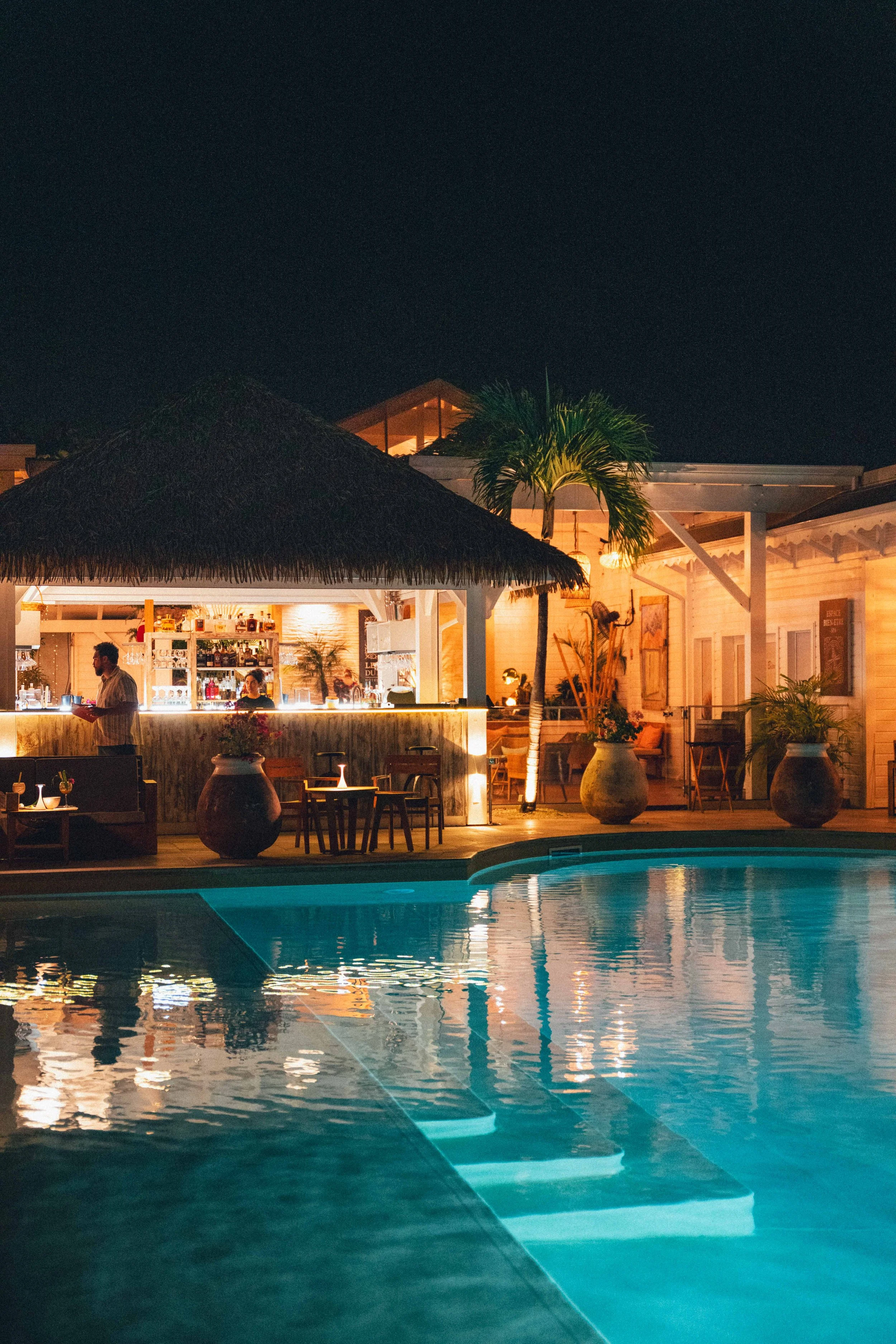 Une piscine éclairée la nuit, entourée d'un espace de détente avec bar, meubles en bois et plantes tropicales, sous un ciel noir.