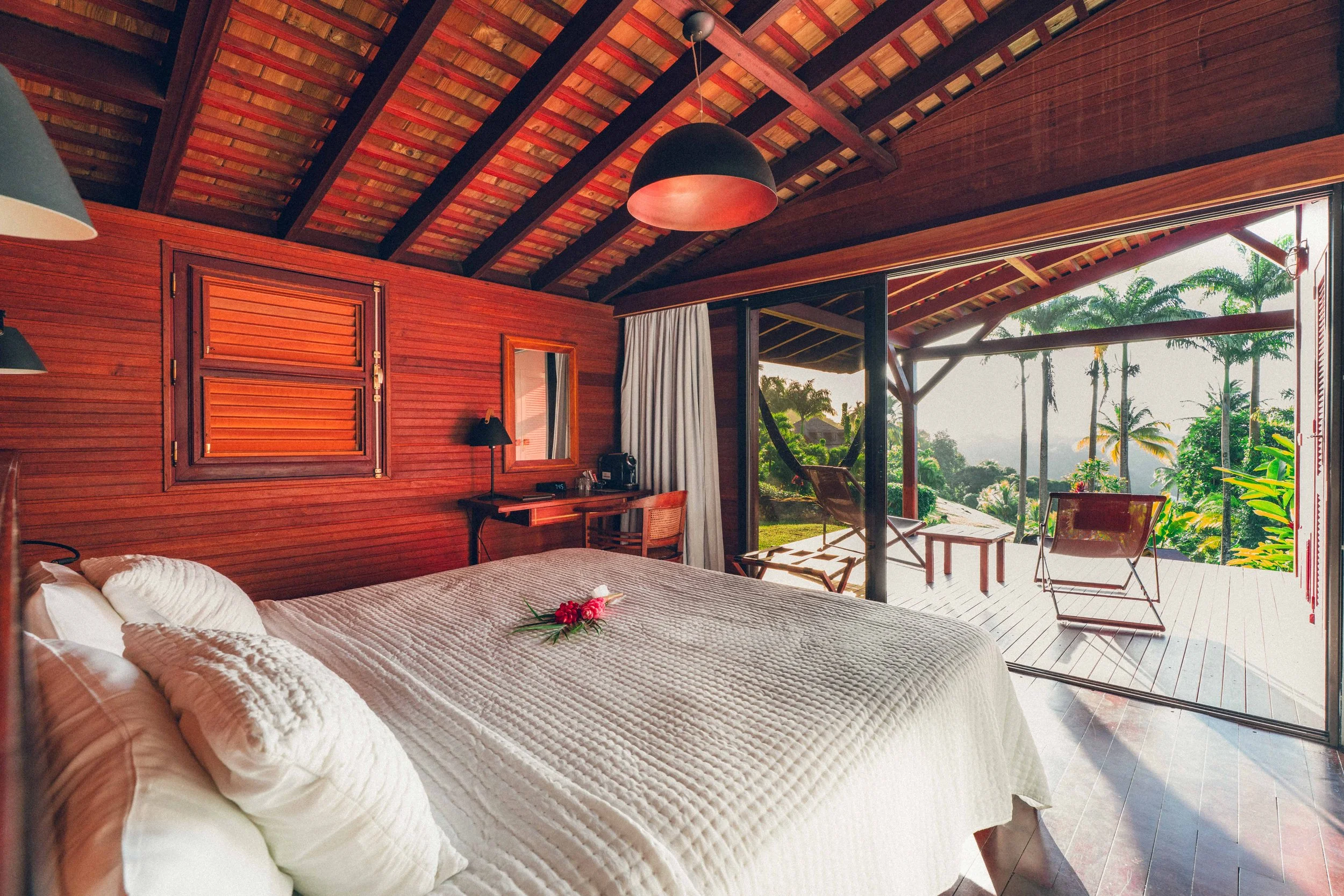 Chambre en bois avec lit, vue ouverte sur une terrasse avec chaises en plein air et végétation tropicale.
