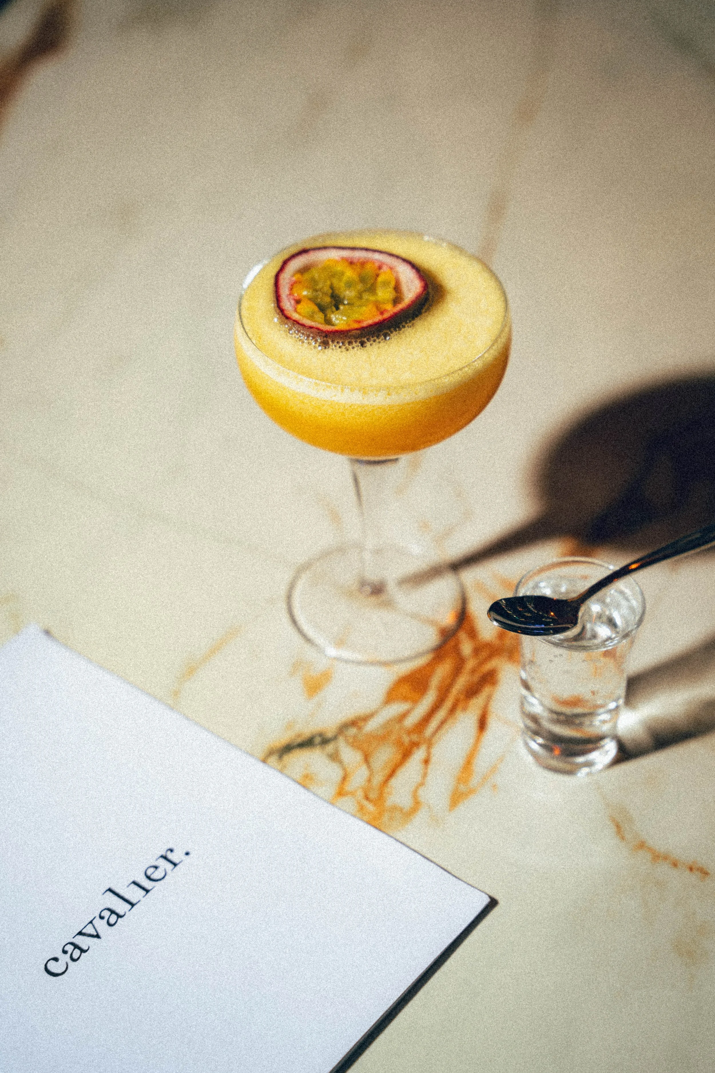 Un verre de cocktail orange décoré avec une tranche de fruit passion sur le dessus, placé sur une table avec une serviette et une cuillère.