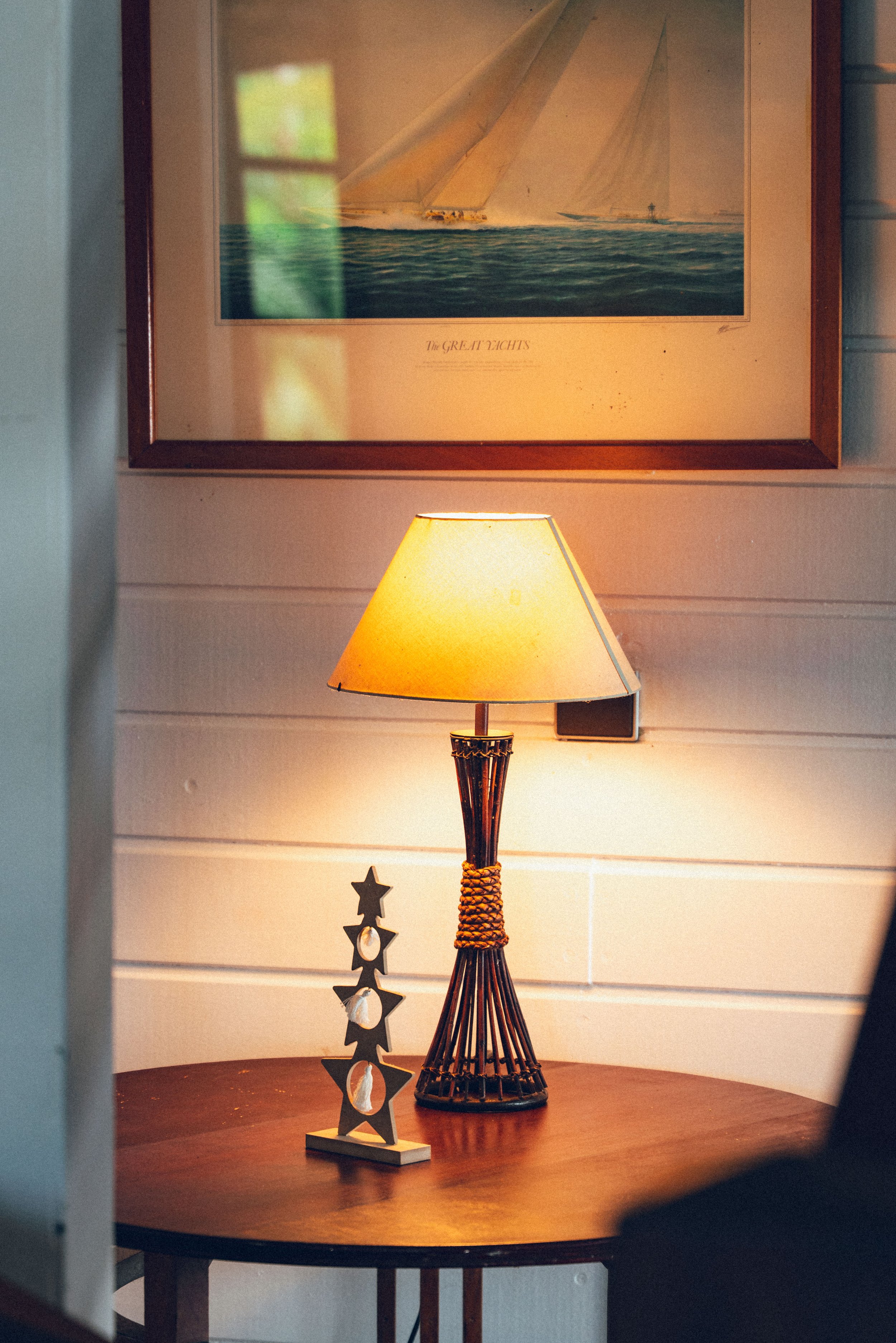 Lampe de table en osier au centre d'une table en bois, avec un tableau représentant une voile de bateau dans le fond. Décorations en forme d'étoiles en bois sur la table.