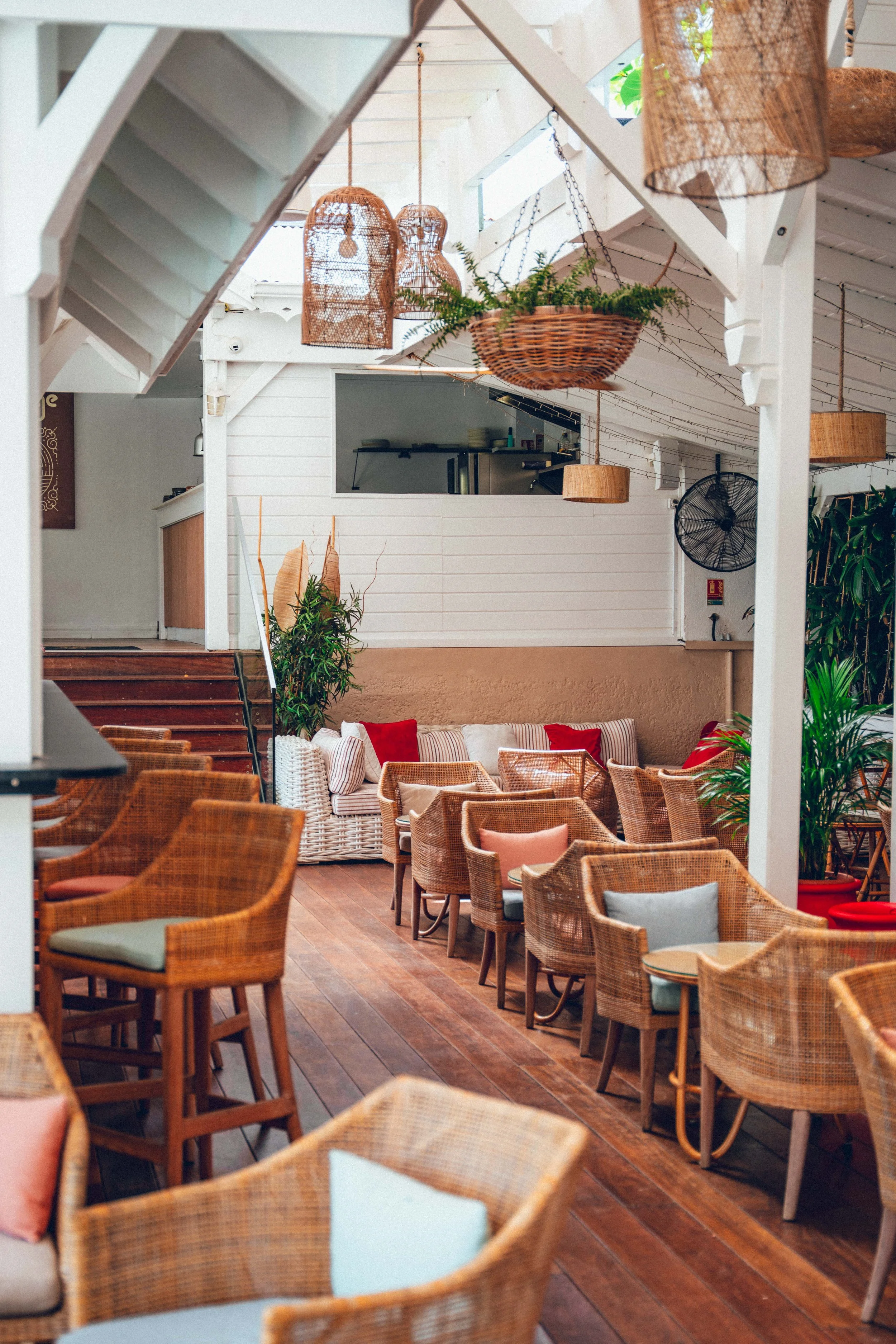 Intérieur d'un restaurant ou d'un lounge avec des chaises en rotin, des coussins, des plantes vertes et des luminaires en rotin suspendus.