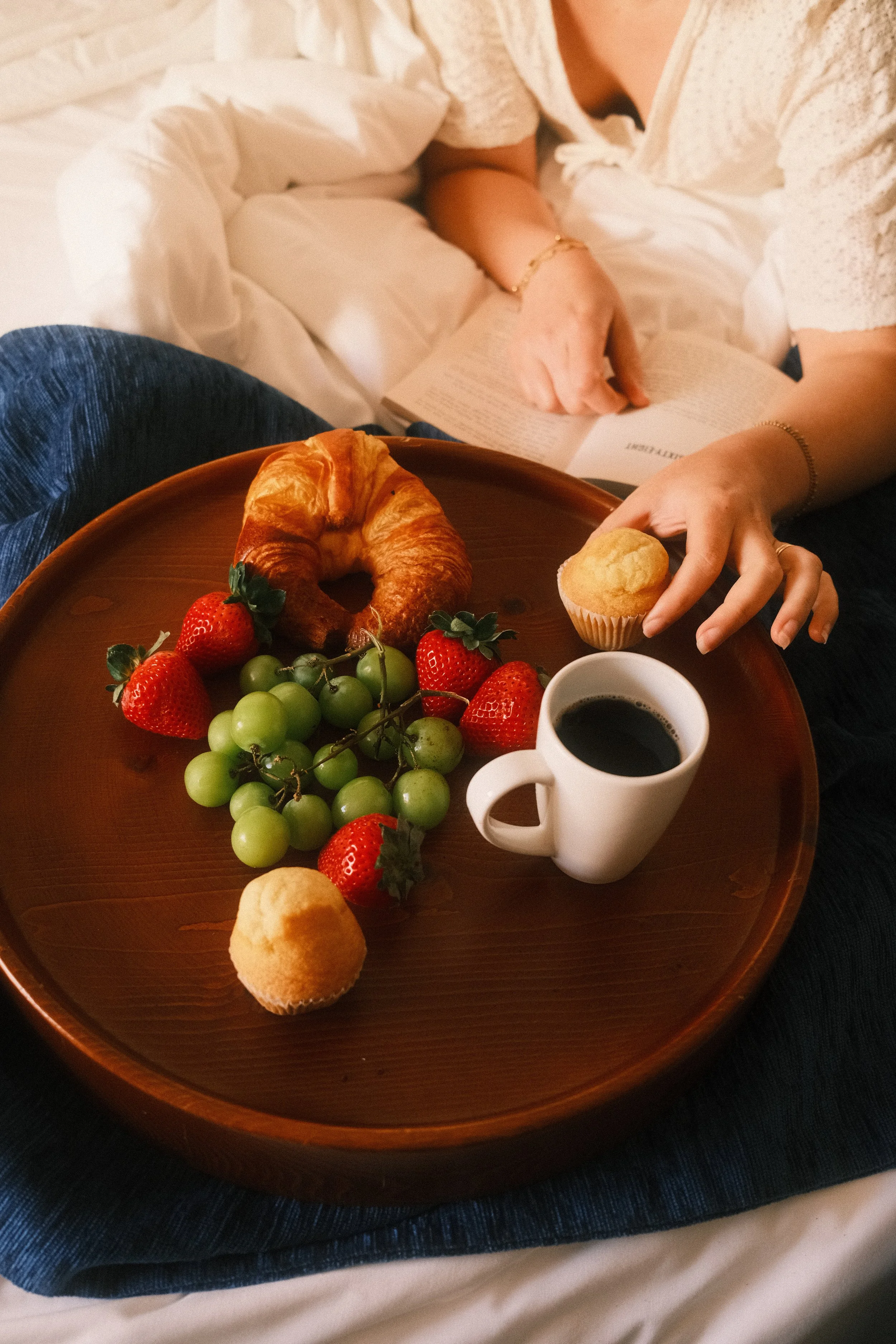 Une personne lit un livre dans un lit avec un plateau contenant un croissant, des fraises, des raisins verts, un muffin, et une tasse de café.