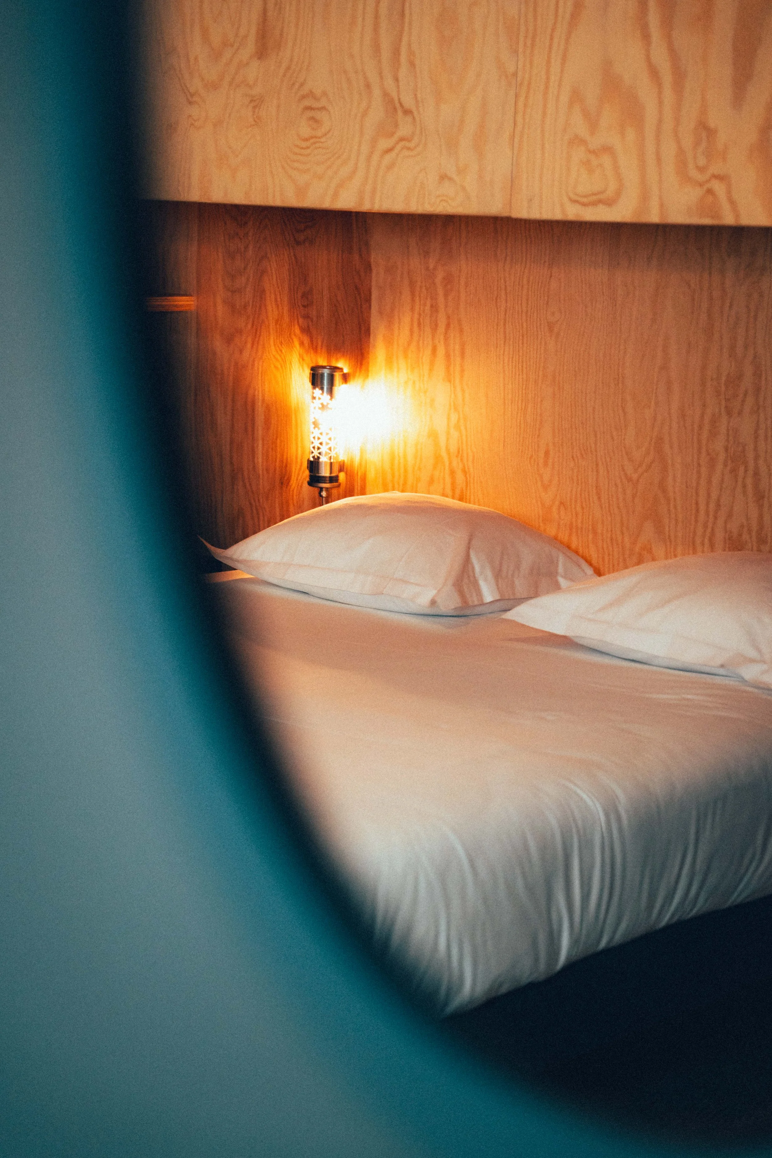 Lumière de lecture allumée sur une tête de lit en bois dans une chambre avec des lits faits avec des draps blancs.