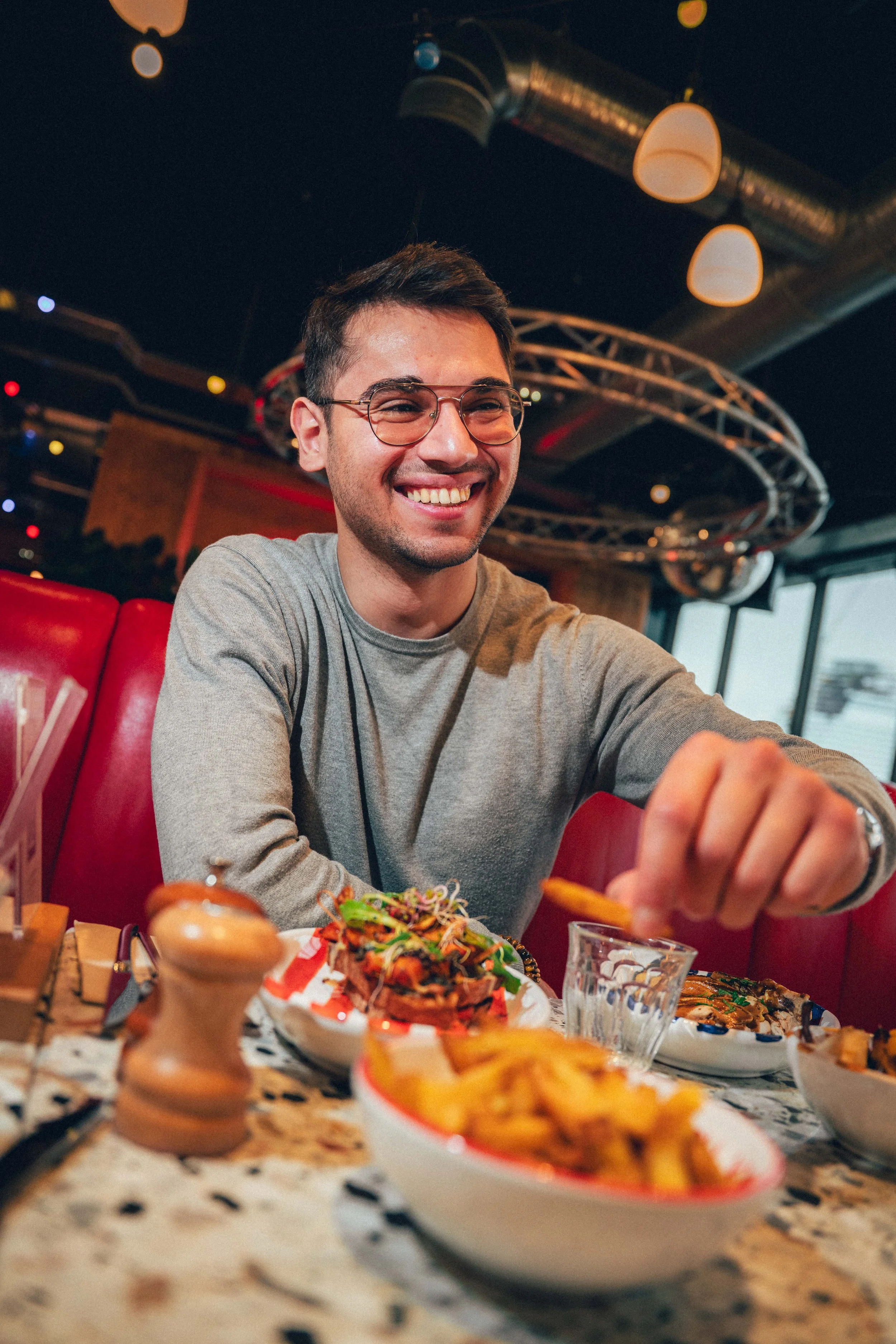 Jeune homme souriant assis à une table de restaurant avec une assiette de nourriture, y compris une salade, des frites et d'autres plats, dans un environnement intérieur moderne avec des lumières suspendues et un plafond noir.