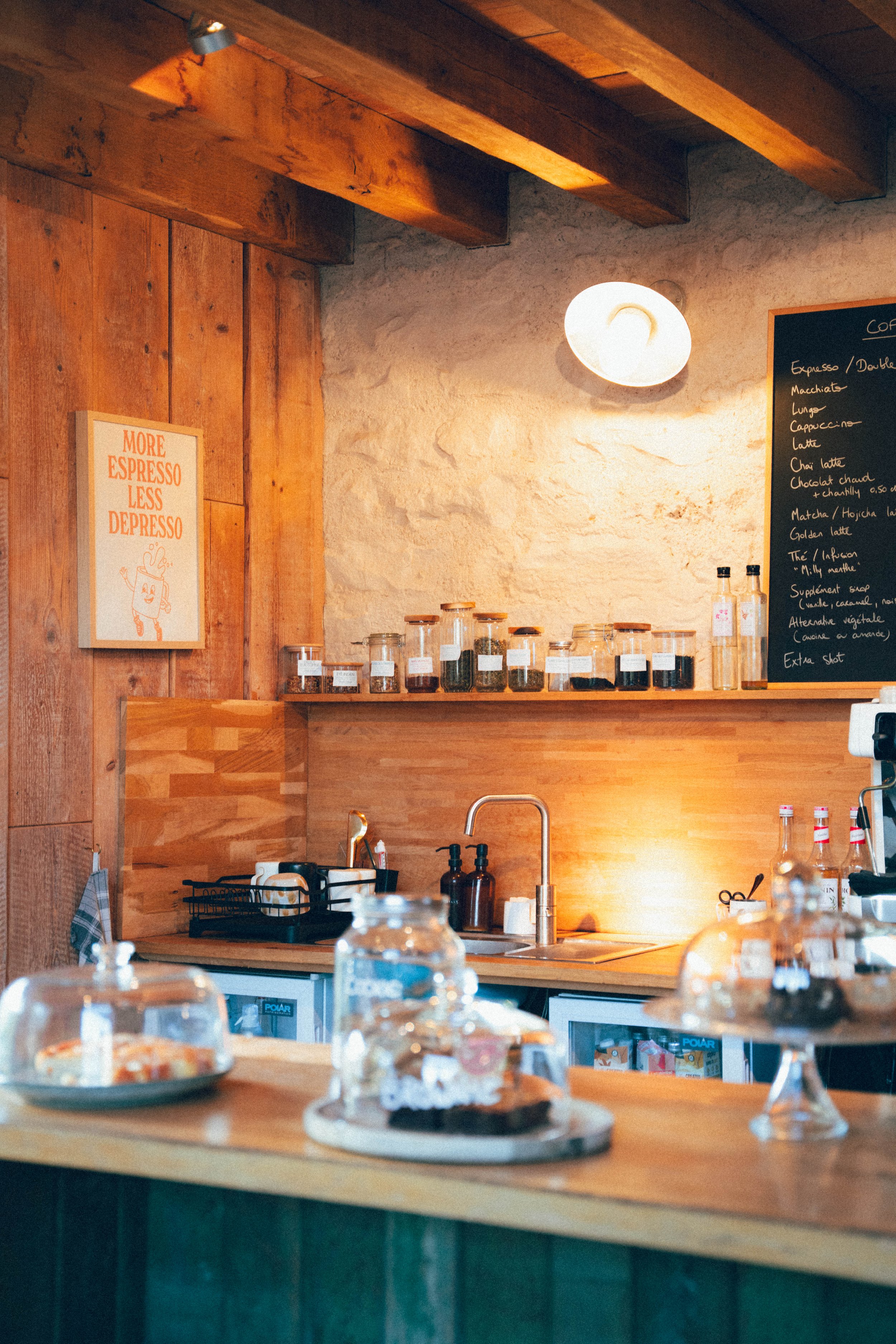 Une devanture de café ou pâtisserie, avec un comptoir en bois, des plats de desserts sous cloches en verre, des bouteilles d'eau, et un tableau noir avec la liste des boissons, dans une ambiance chaleureuse en bois.
