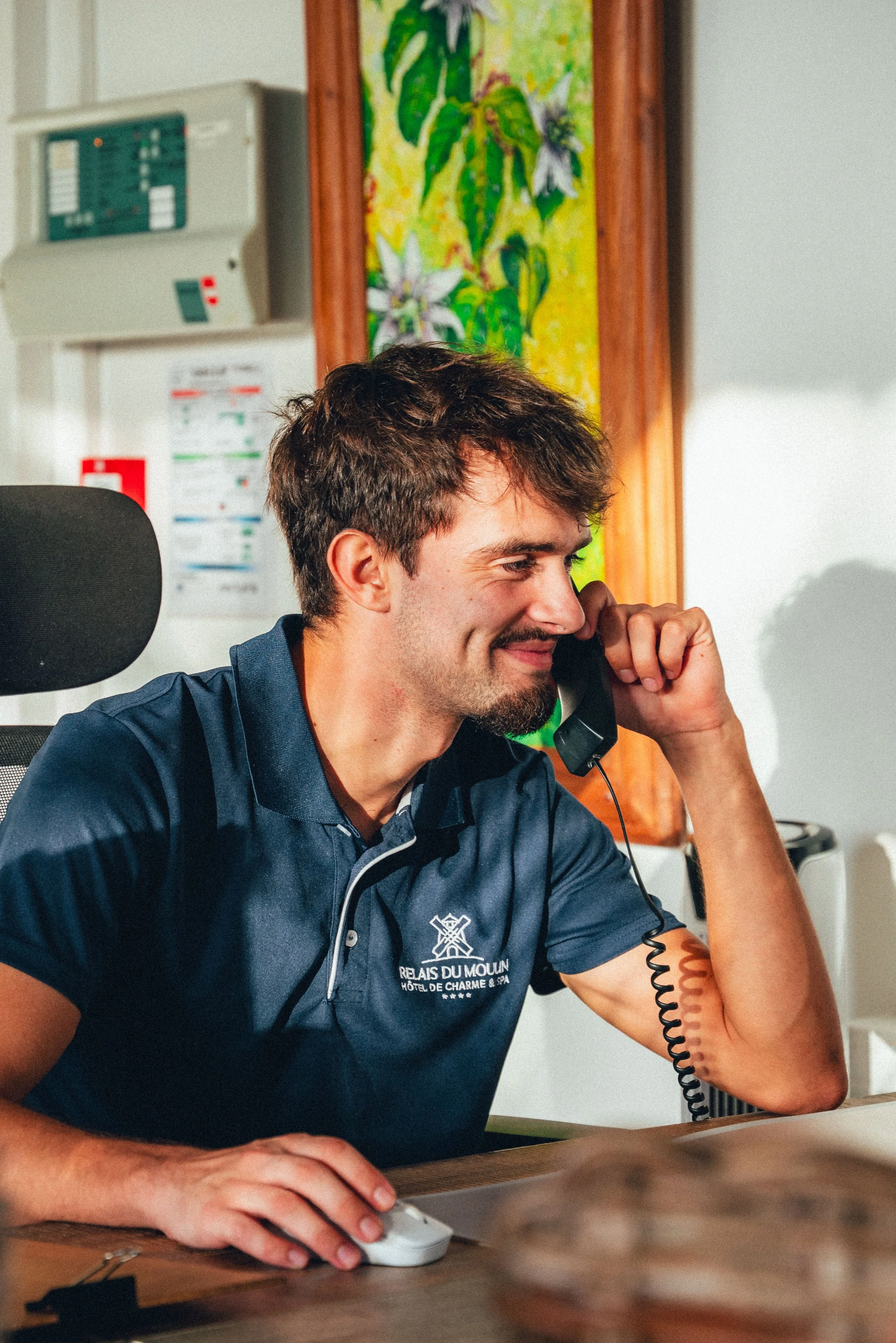 Jeune homme souriant au téléphone, portant un polo bleu marine avec logo de l'hôtel, assis à un bureau.