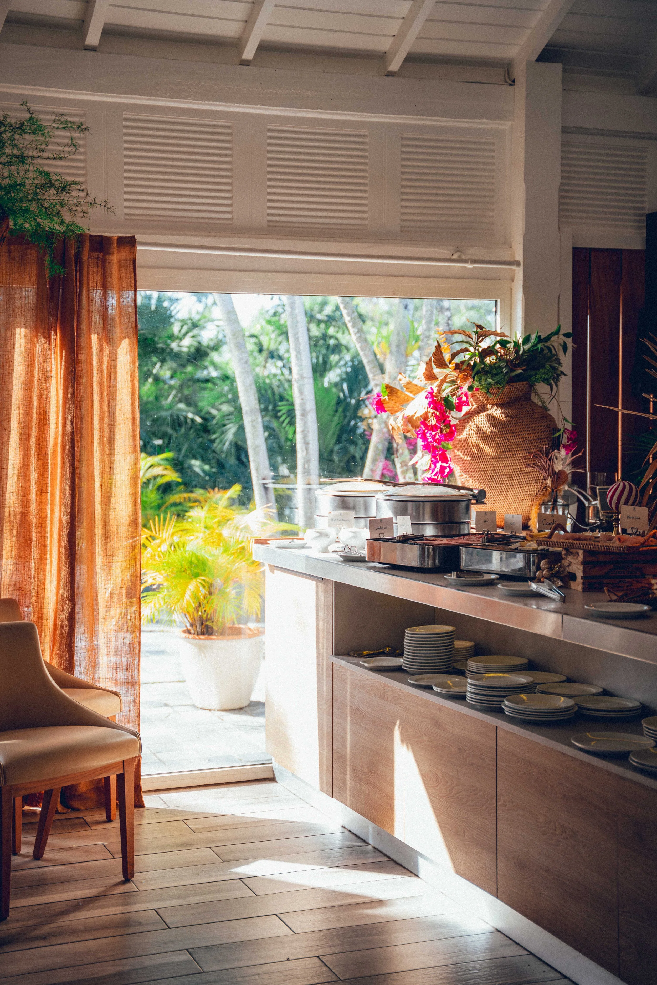 Intérieur d'un restaurant ou d'une cafétéria avec comptoir de service, vaisselle empilée, plantes décoratives, grande fenêtre laissant entrer la lumière naturelle, et vue sur un jardin extérieur avec des arbres et des plantes tropicales.