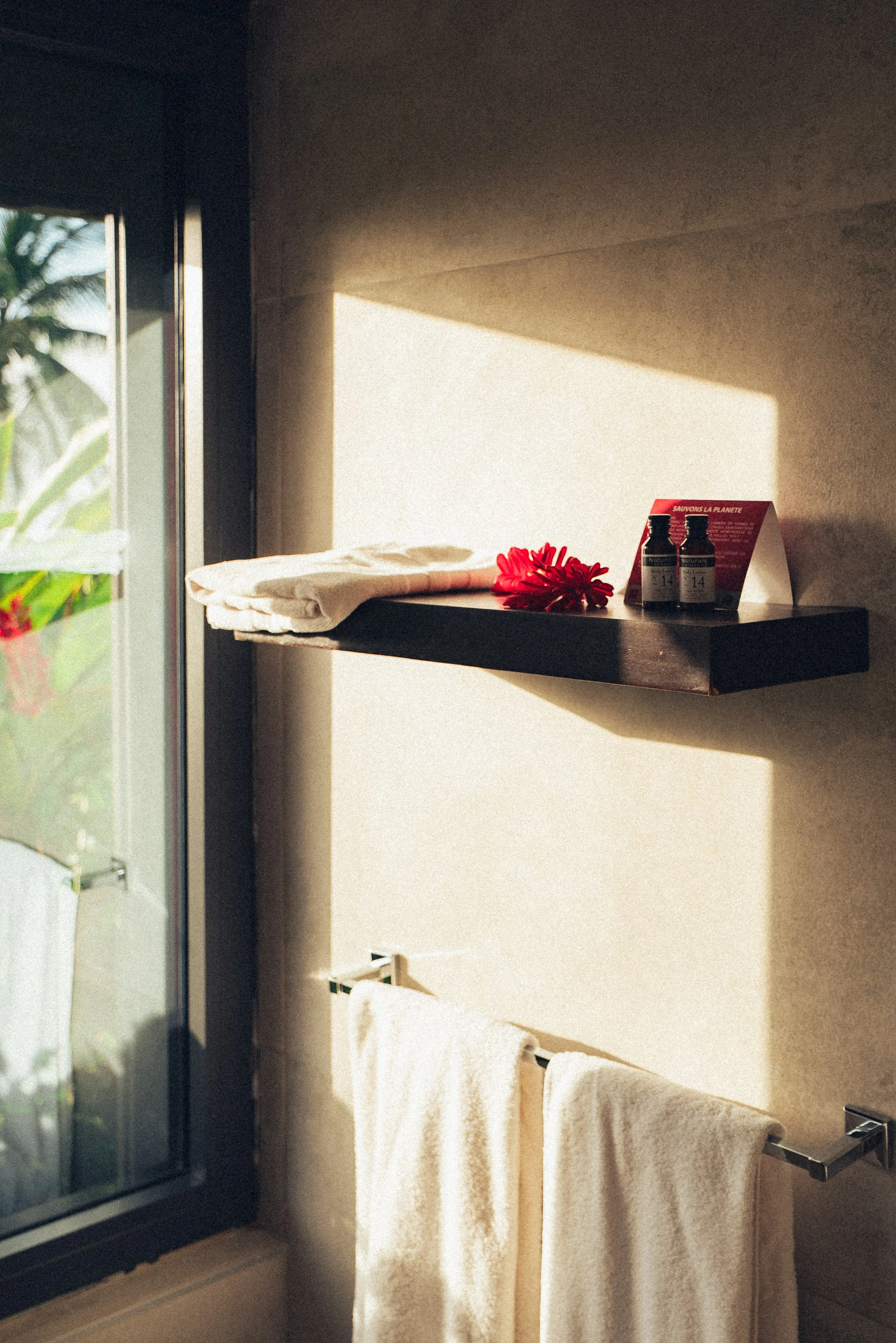 Une étagère noire fixée au mur avec deux bouteilles et une affiche promotionnelle, une serviette blanche pliée et une serviette beige suspendue, près d'une fenêtre laissant passer la lumière