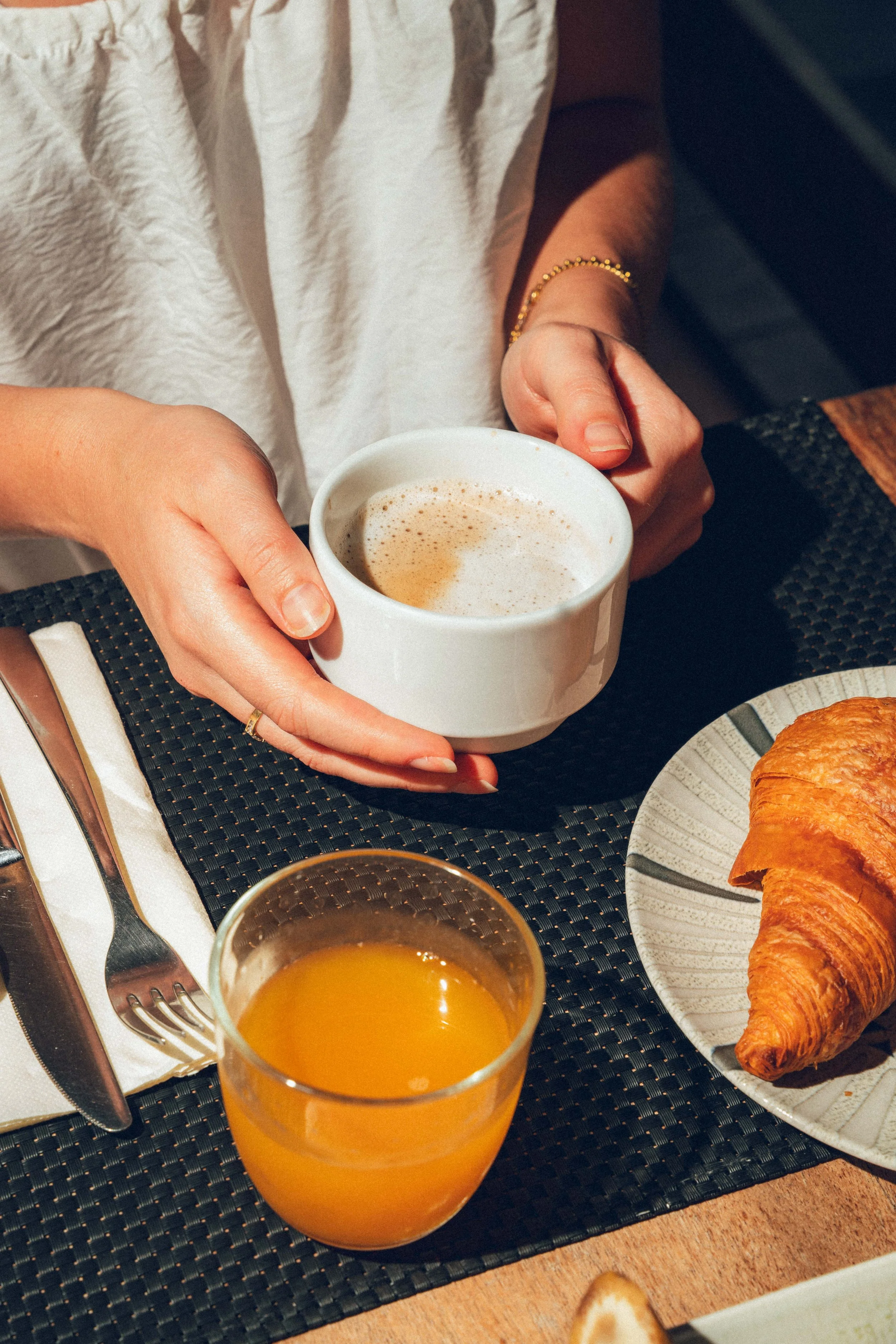 Une personne tient une tasse de café ou de cappuccino, avec un croissant sur une assiette, un verre de jus d'orange, et un repas de petit-déjeuner sur une table.