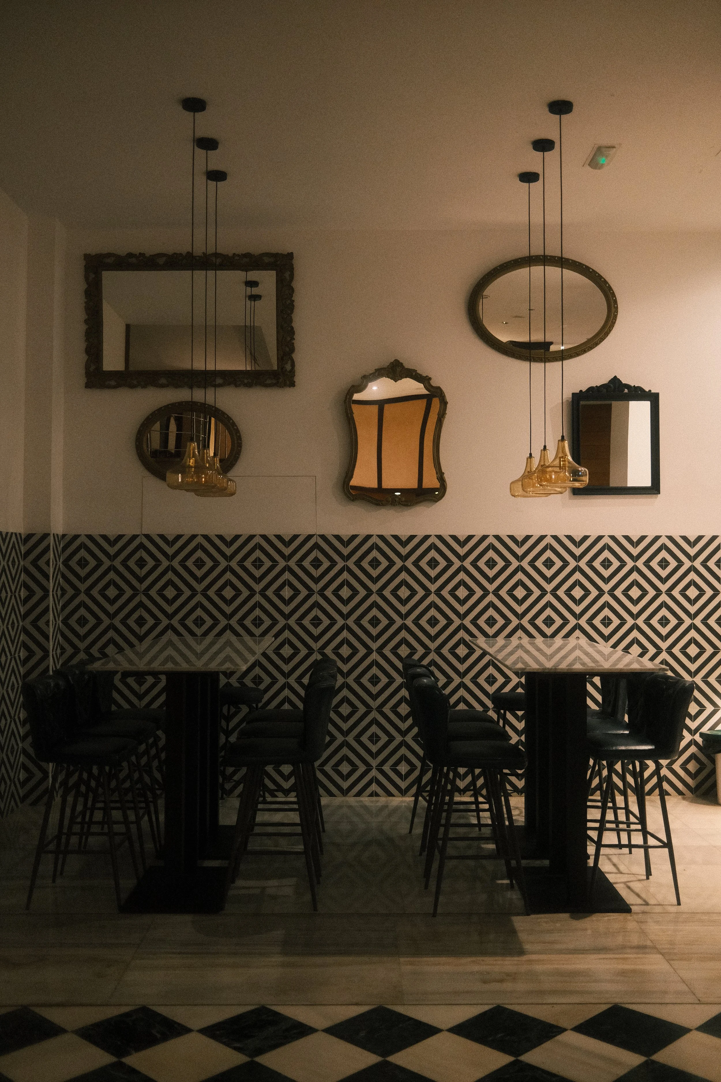 Une salle à manger ou un restaurant avec un mur en diagonale noir et blanc, plusieurs miroirs de styles différents accrochés au mur, des tables rondes et des chaises en métal noir. Plusieurs suspensions lumineuses en verre ambré sont suspendues au pl