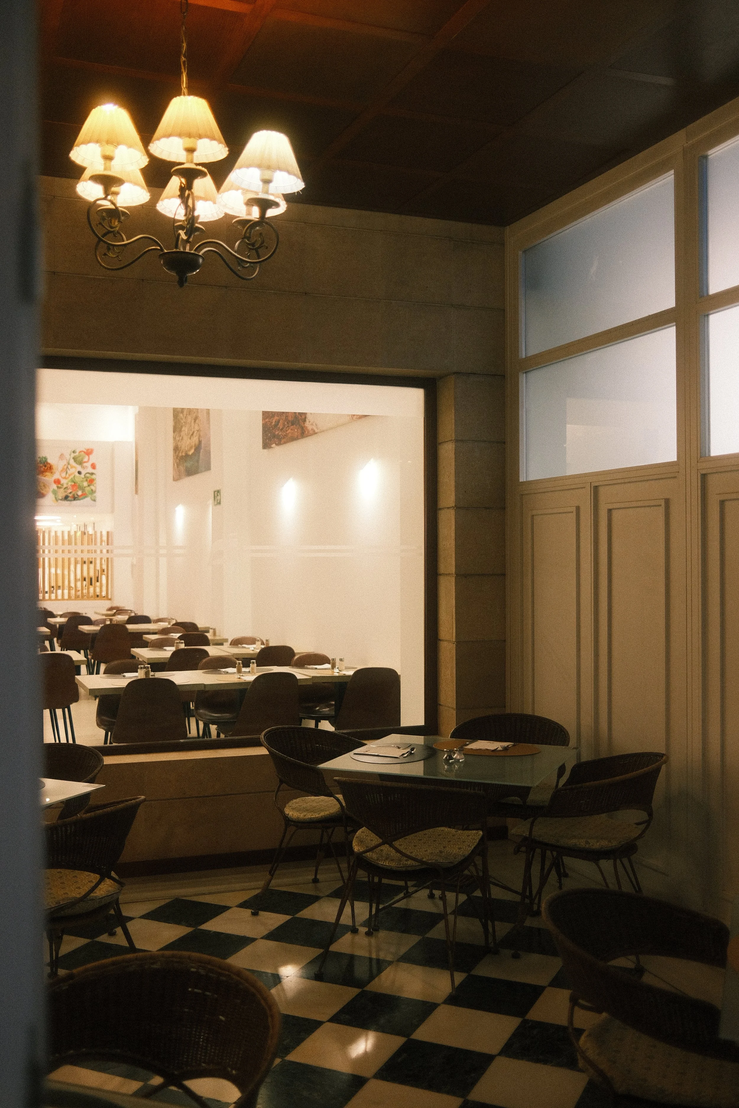 Salle à manger d'un restaurant avec des tables, chaises, un mur en verre, et un plafond en bois. Un lustre avec des abat-jours donne une lumière chaleureuse.