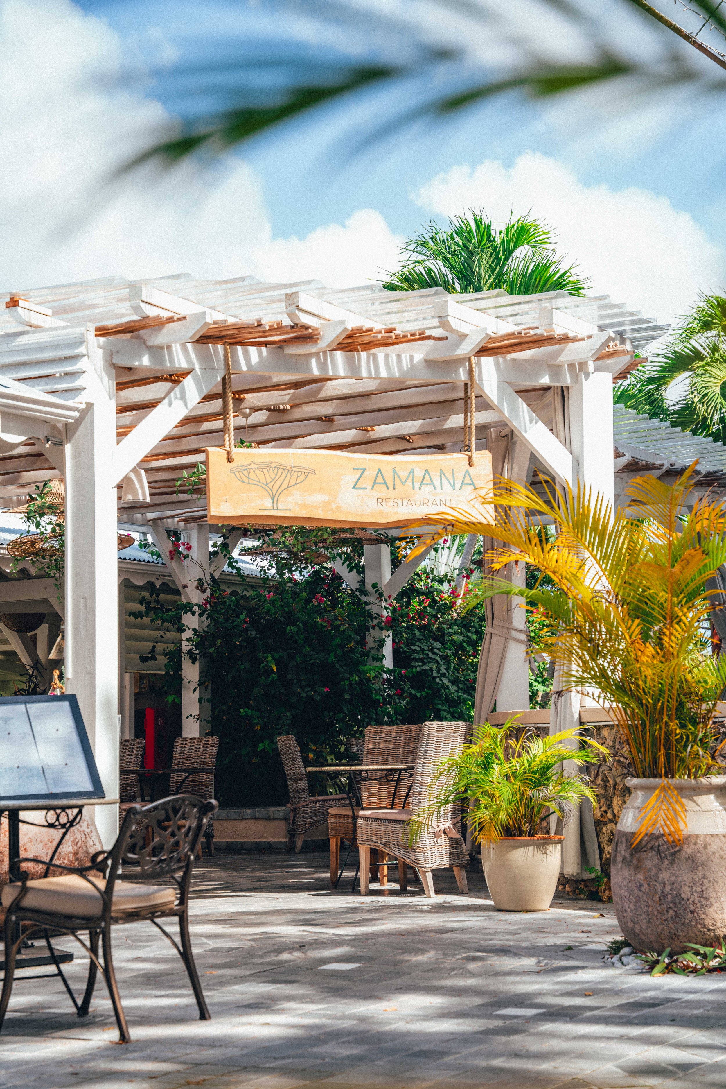Une terrasse extérieure d'un restaurant avec des chaises en rotin, des tables, des plantes en pot et un panneau en bois avec le nom 'ZAMANA RESTAURANT' suspendu.