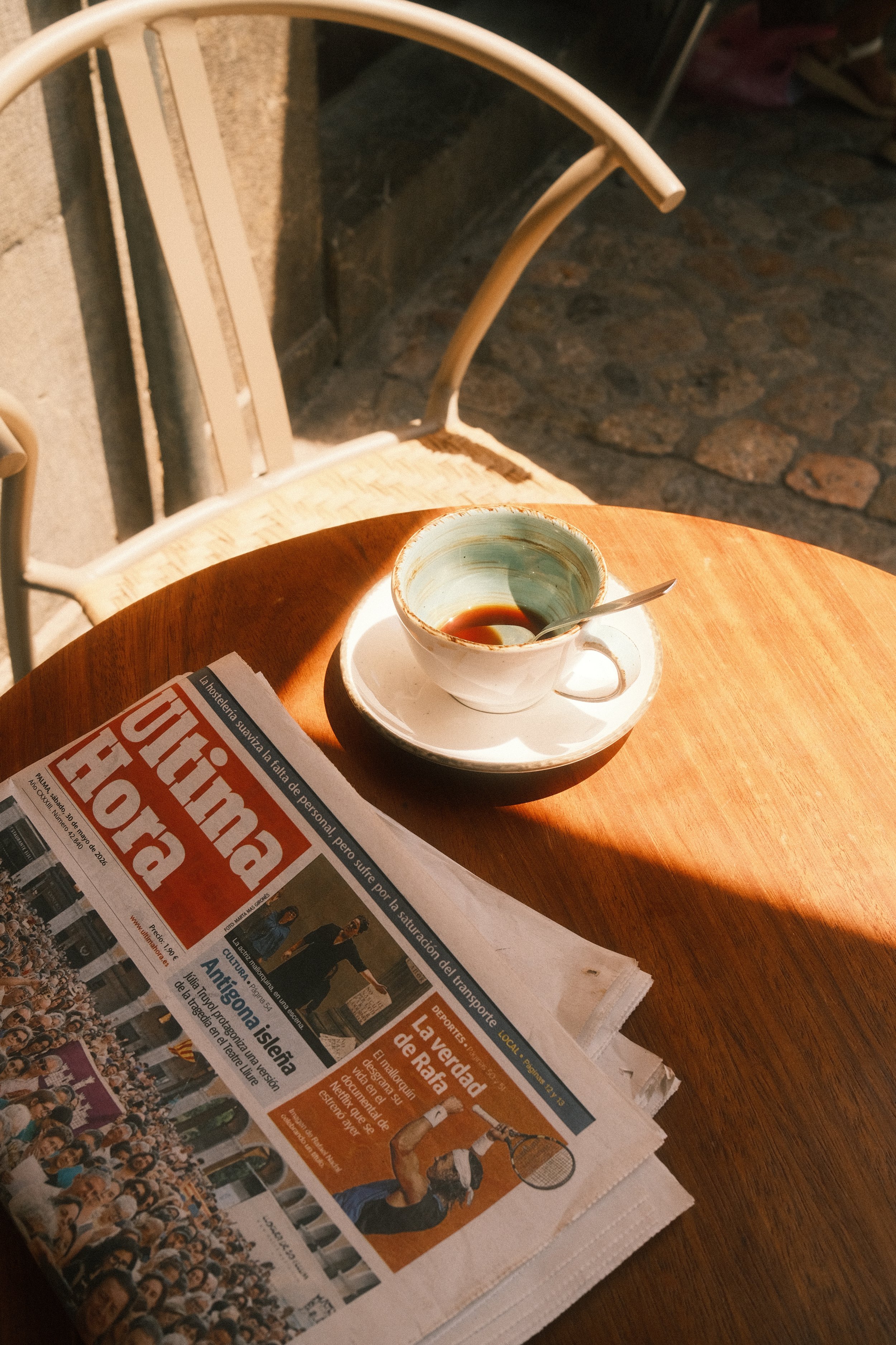 Une tasse de café à moitié pleine sur une assiette blanche, posée sur une table en bois avec un journal à côté, sous un éclairage chaud à l'extérieur.