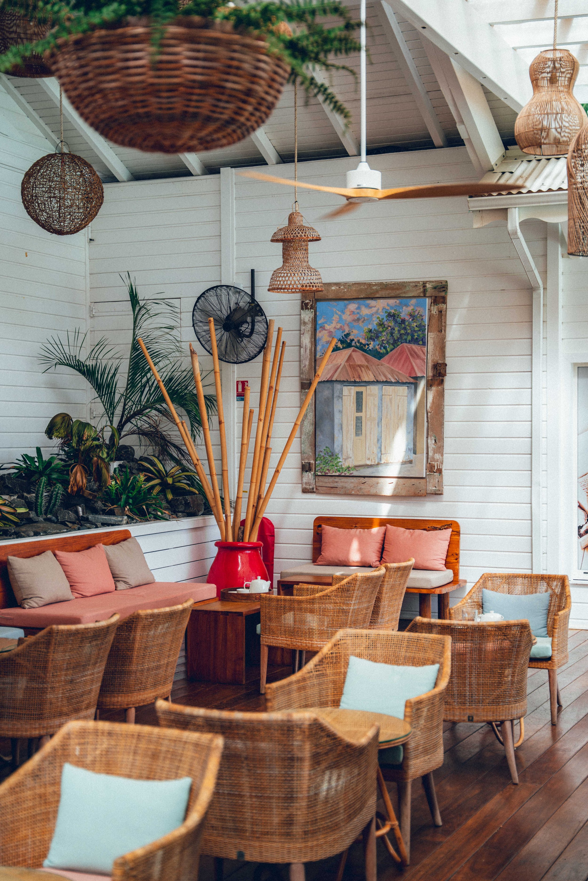 Photographie intérieur d'un restaurant avec des chaises en rotin autour de tables en bois, un canapé avec des coussins, des plantes vertes, des lampes en osier suspendues, un tableau peint de maisons avec des toits en tuiles rouges.